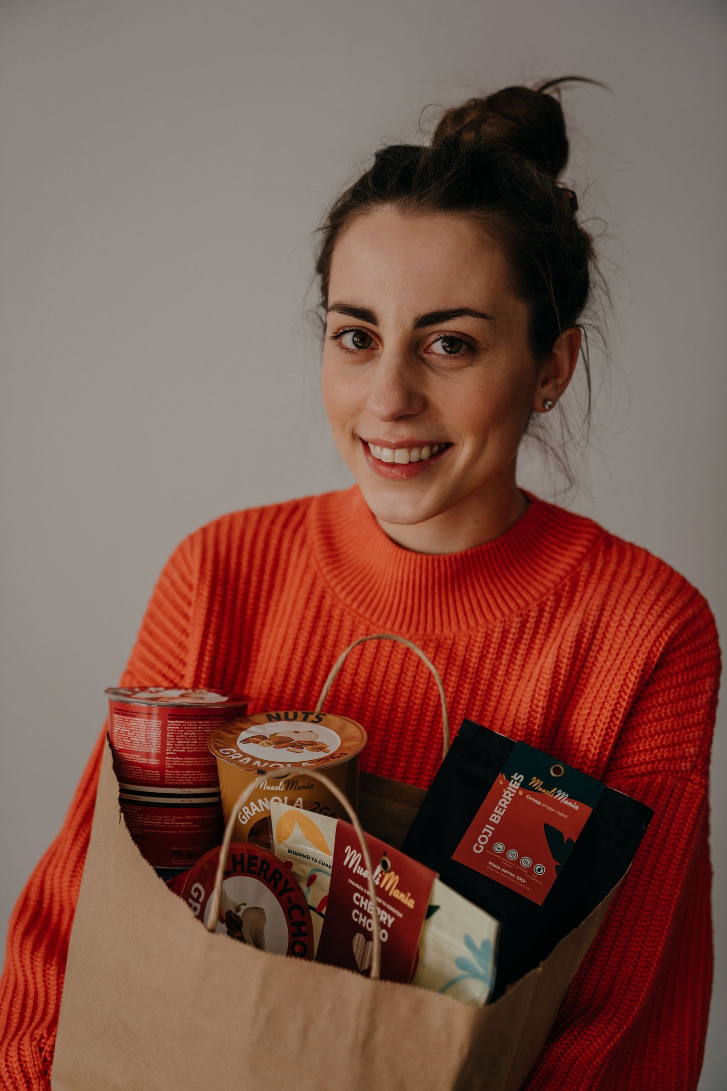 Smiling woman in orange sweater holding a paper bag filled with Muesli Mania granola and food products.