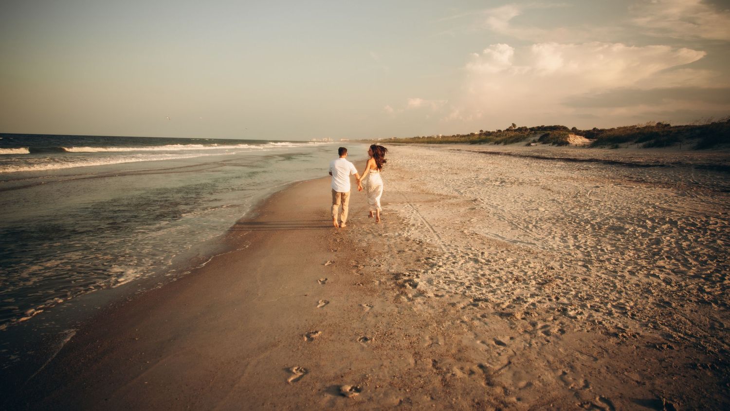 Couple holding hands walking along a sandy beach at sunset with ocean waves and cloudy sky in background.