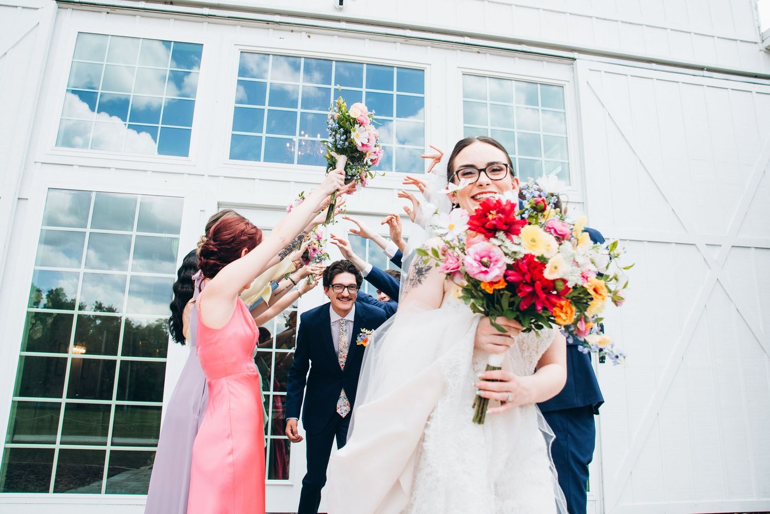Bride in white gown holding colorful bouquet with wedding party celebrating outside a white building.