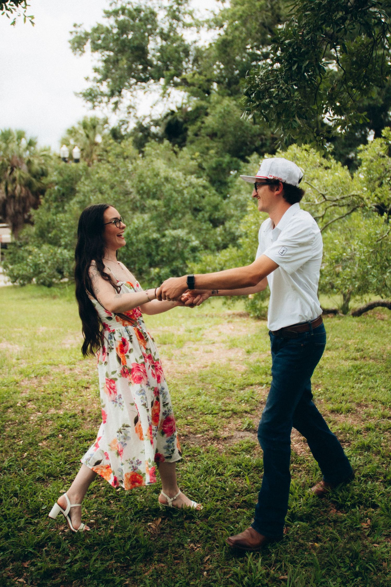 Happy couple holding hands and dancing together in green park, woman wearing floral sundress.