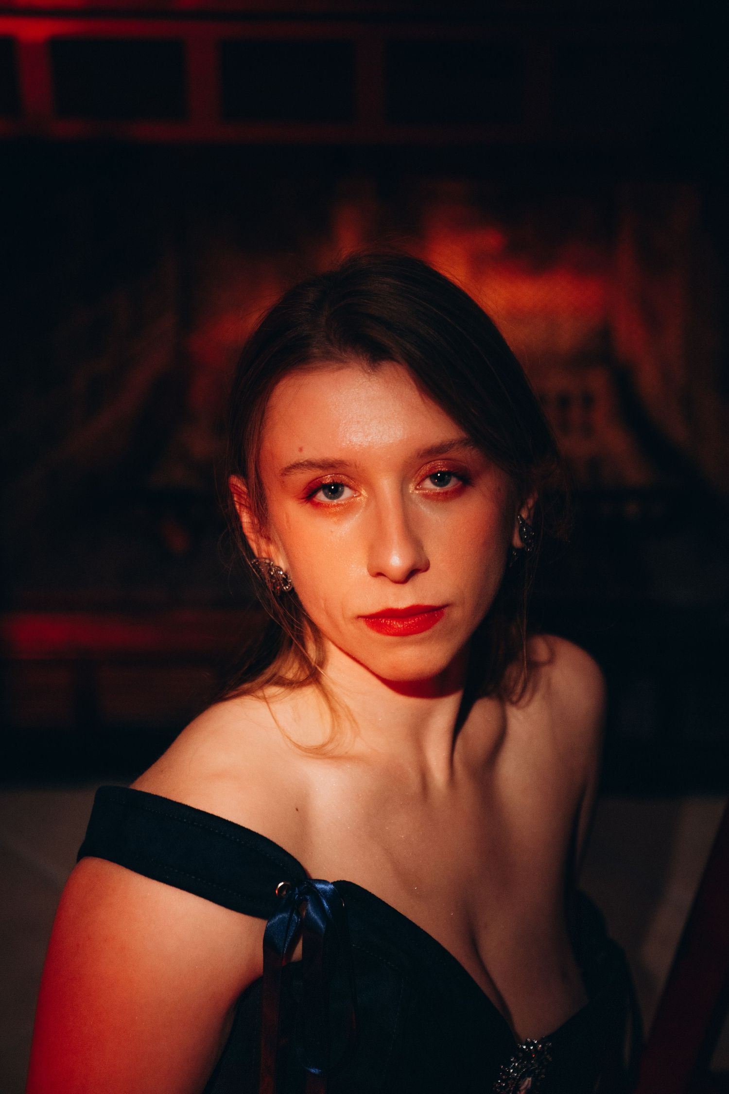 Young woman in off-shoulder black dress with red lips poses by a glowing fireplace in dramatic lighting.