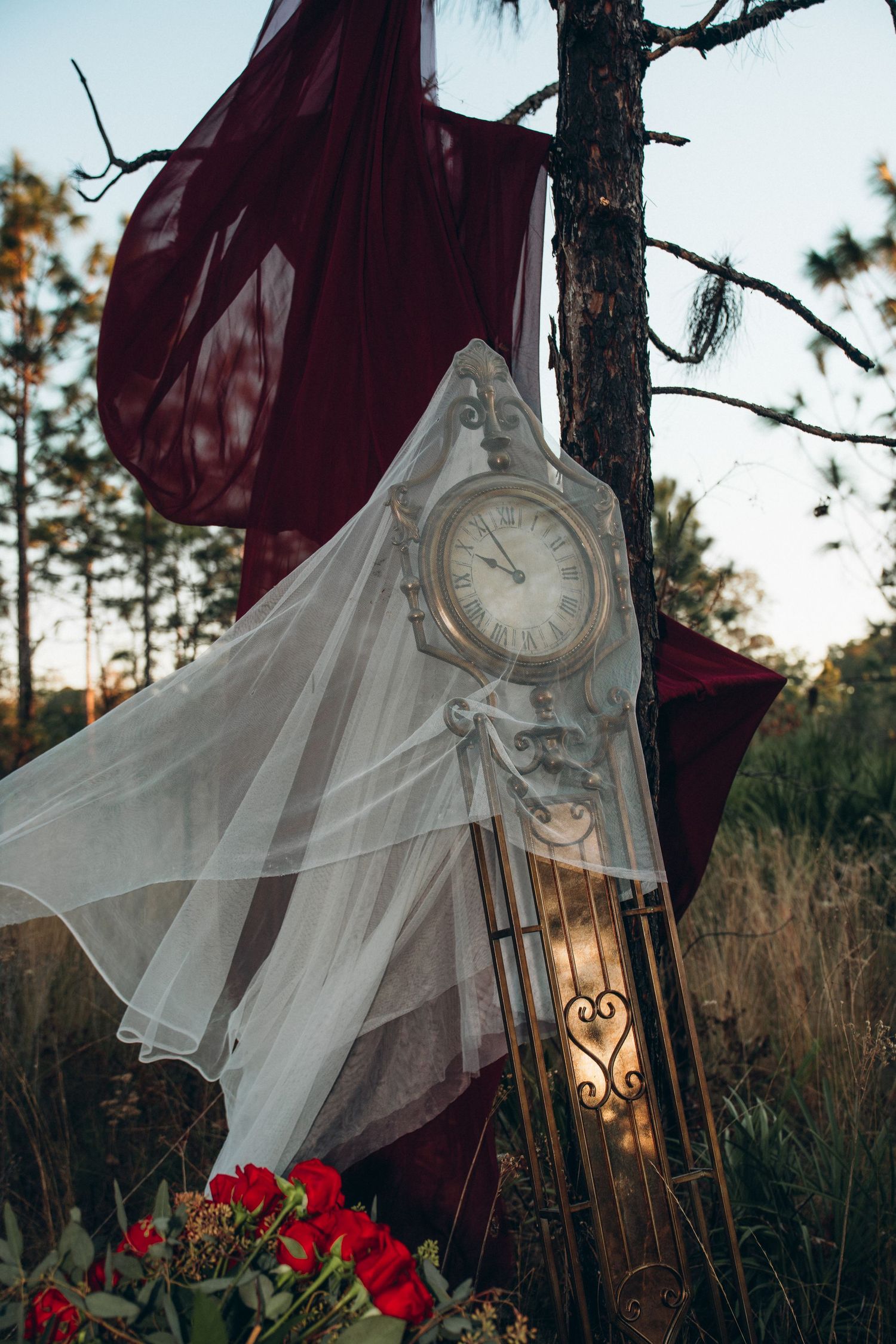 Vintage ornate clock draped in white tulle with red fabric and flowers in a mystical forest setting.