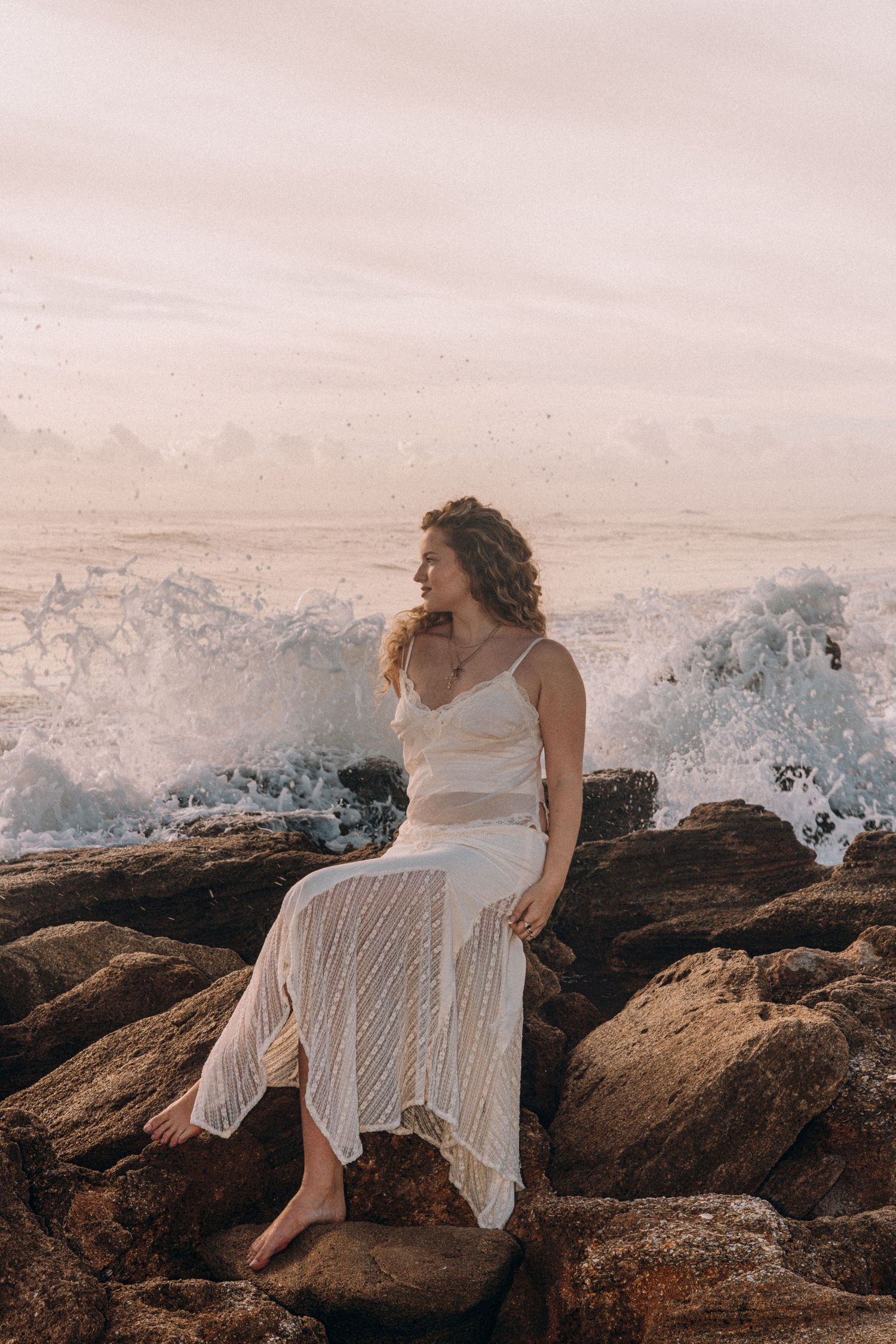 Woman in white lace dress sitting on rocky shore with crashing waves at golden hour.