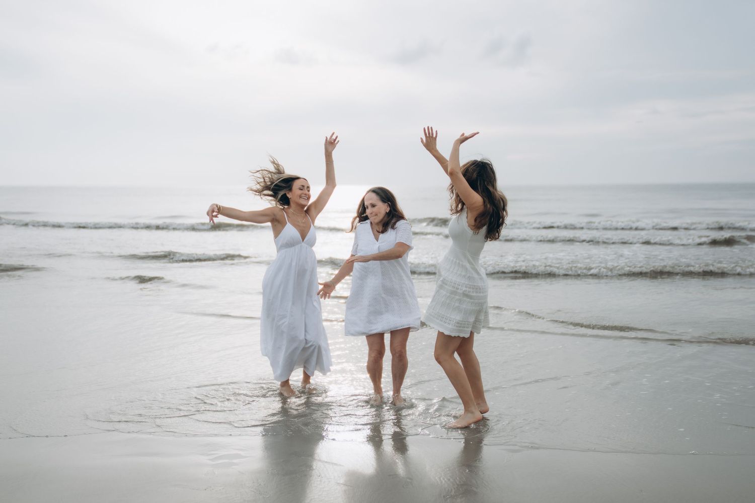 Three women in white dresses dancing joyfully on a beach with ocean waves in the background.