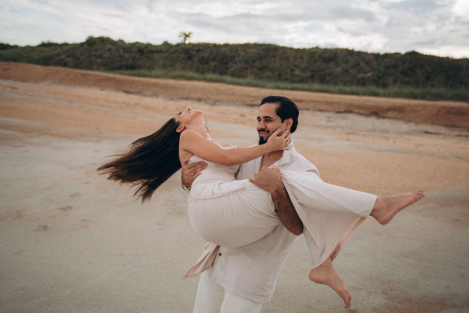 Couple in white outfits embracing on a sandy beach with dramatic clouds.