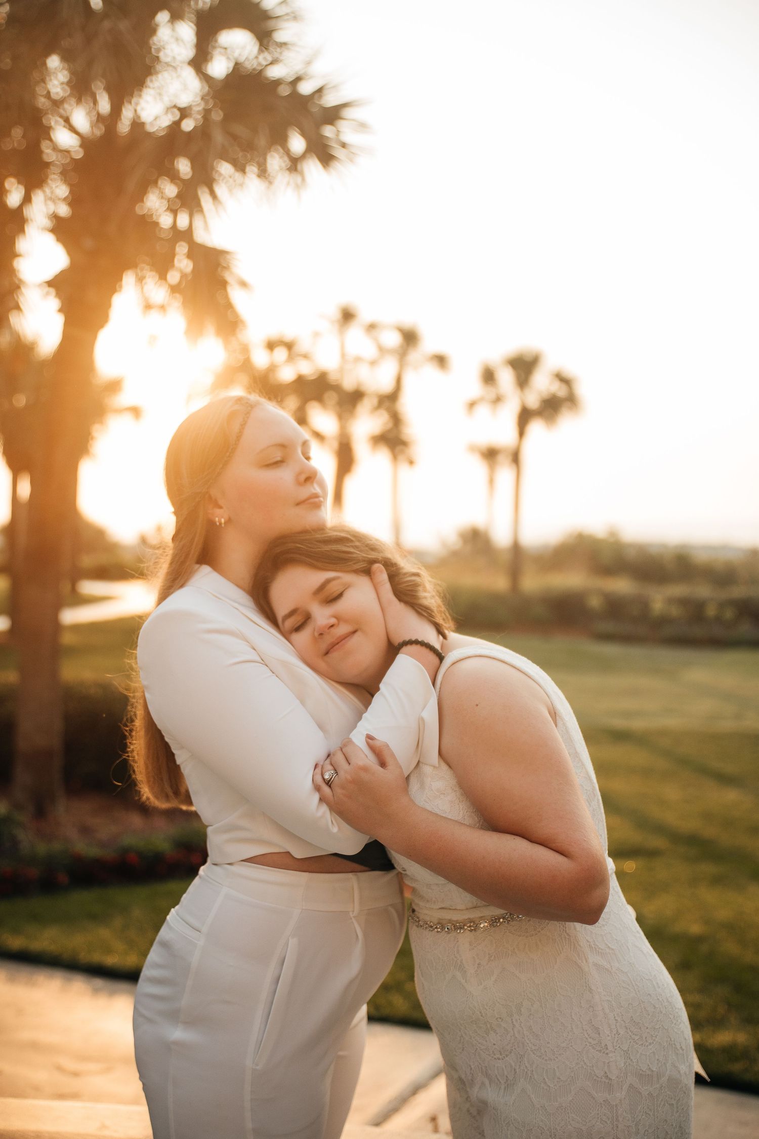 Two people share an intimate moment in white attire against a warm sunset and palm tree backdrop.