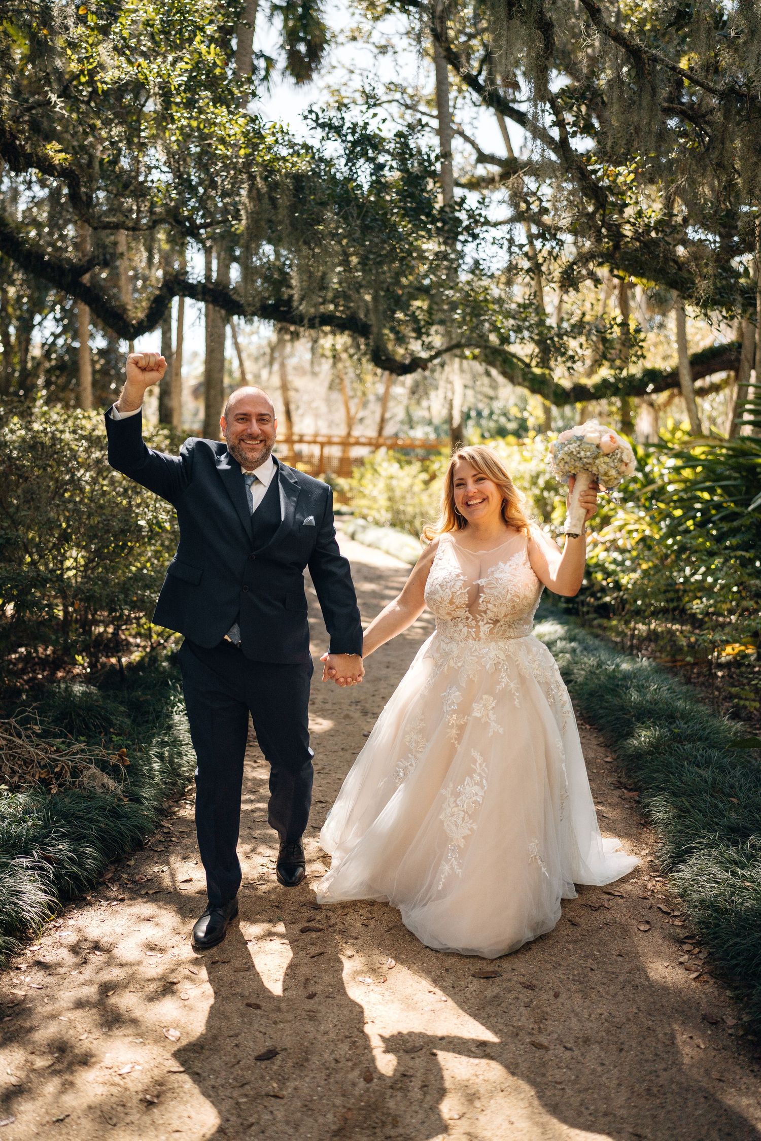 Newlyweds share joyful moments while walking along a garden path under Spanish moss.