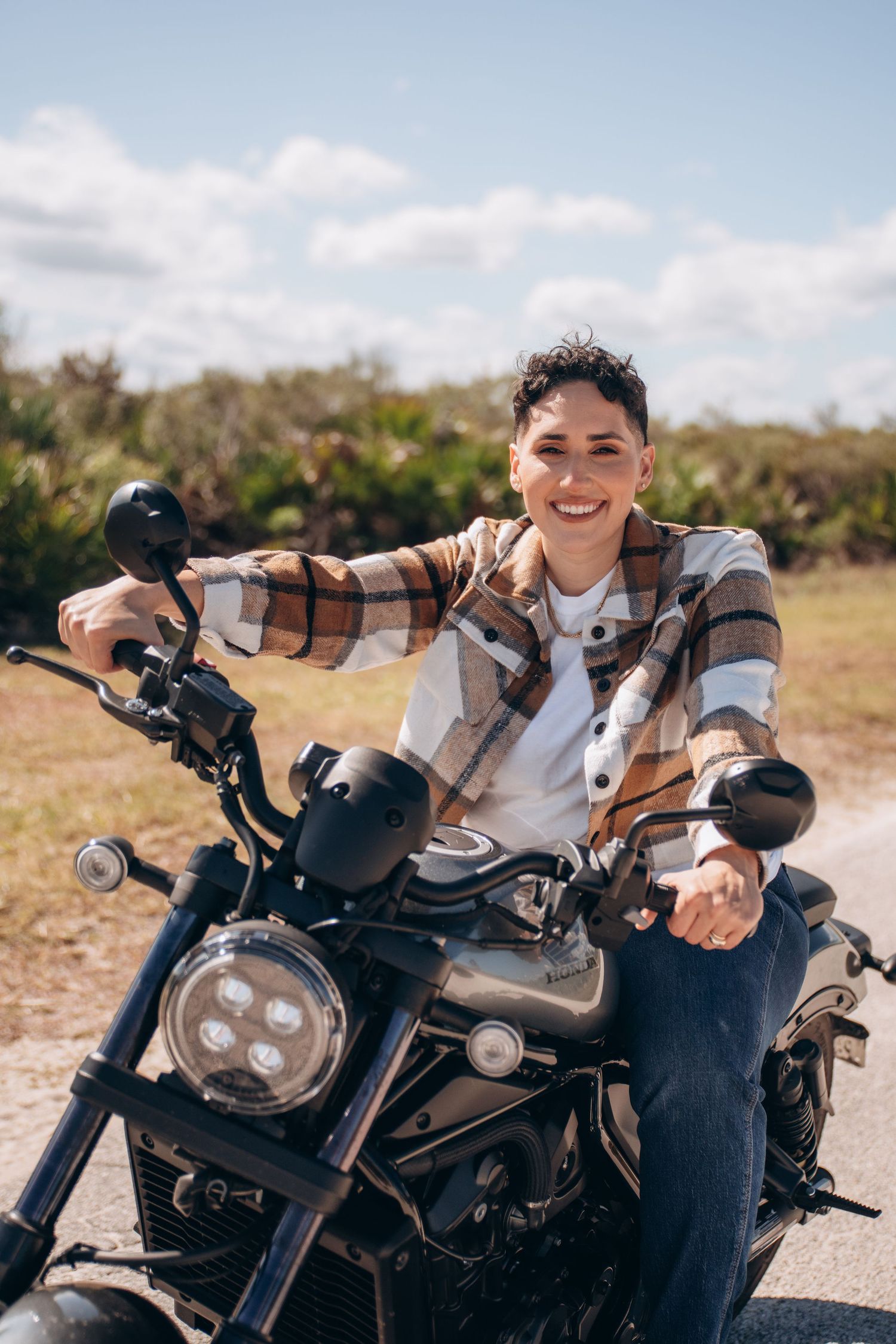 A motorcyclist in casual attire sits on a black motorcycle on a sunny day in a natural outdoor setting.