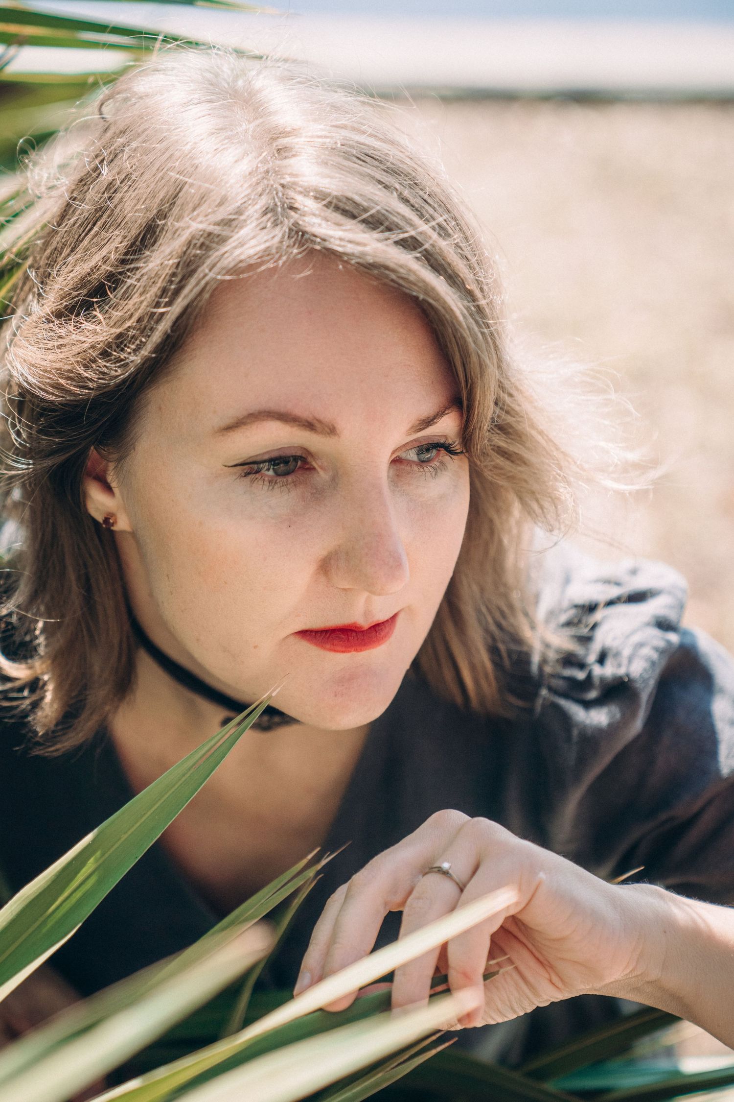 An artistic portrait through palm fronds showing a woman wearing red lipstick and a black choker.