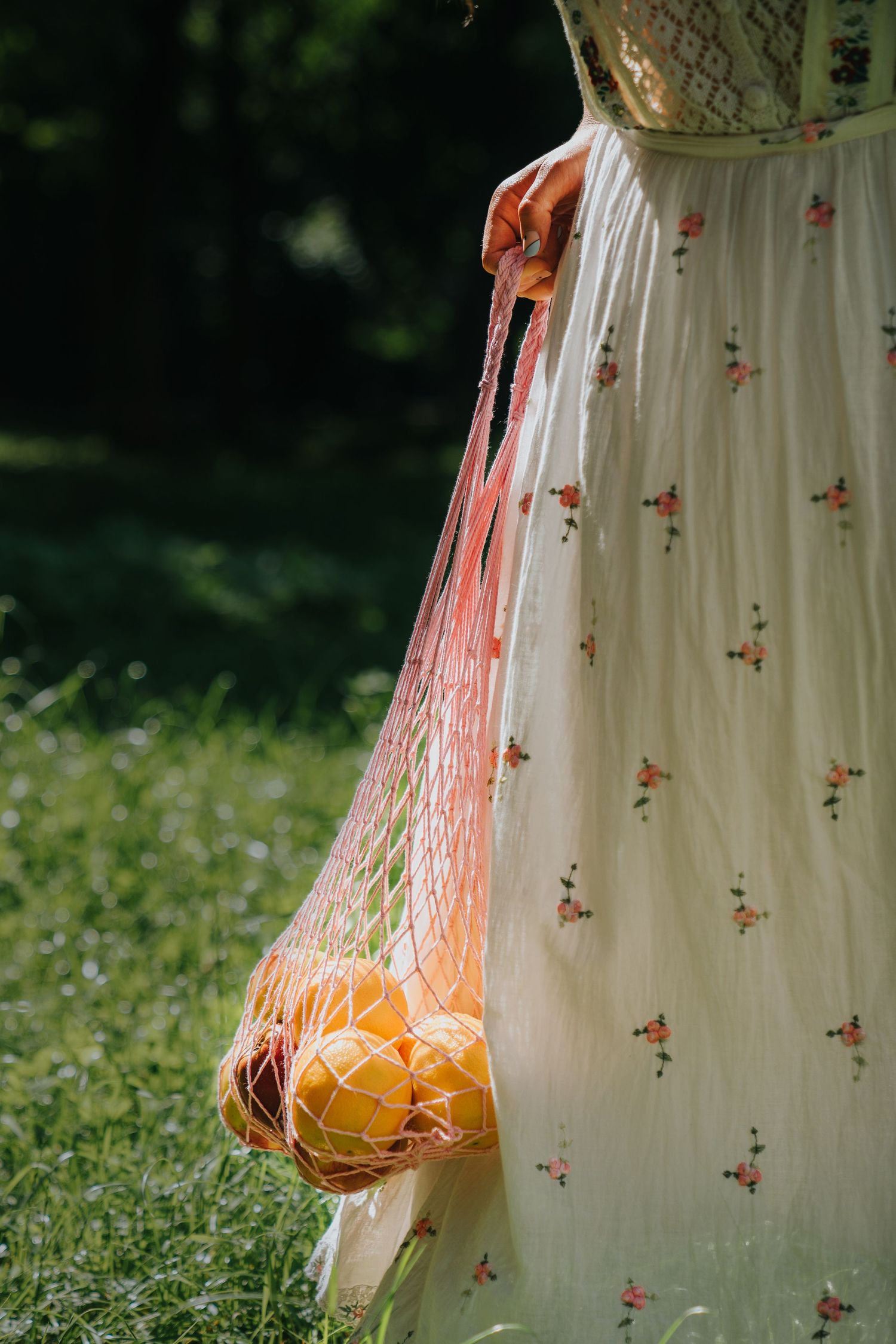 Woman in floral dress carrying a mesh bag filled with fresh oranges in a sunny green garden.