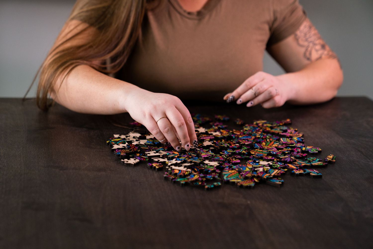 Woman with tattoo assembling colorful jigsaw puzzle pieces on dark wooden table.