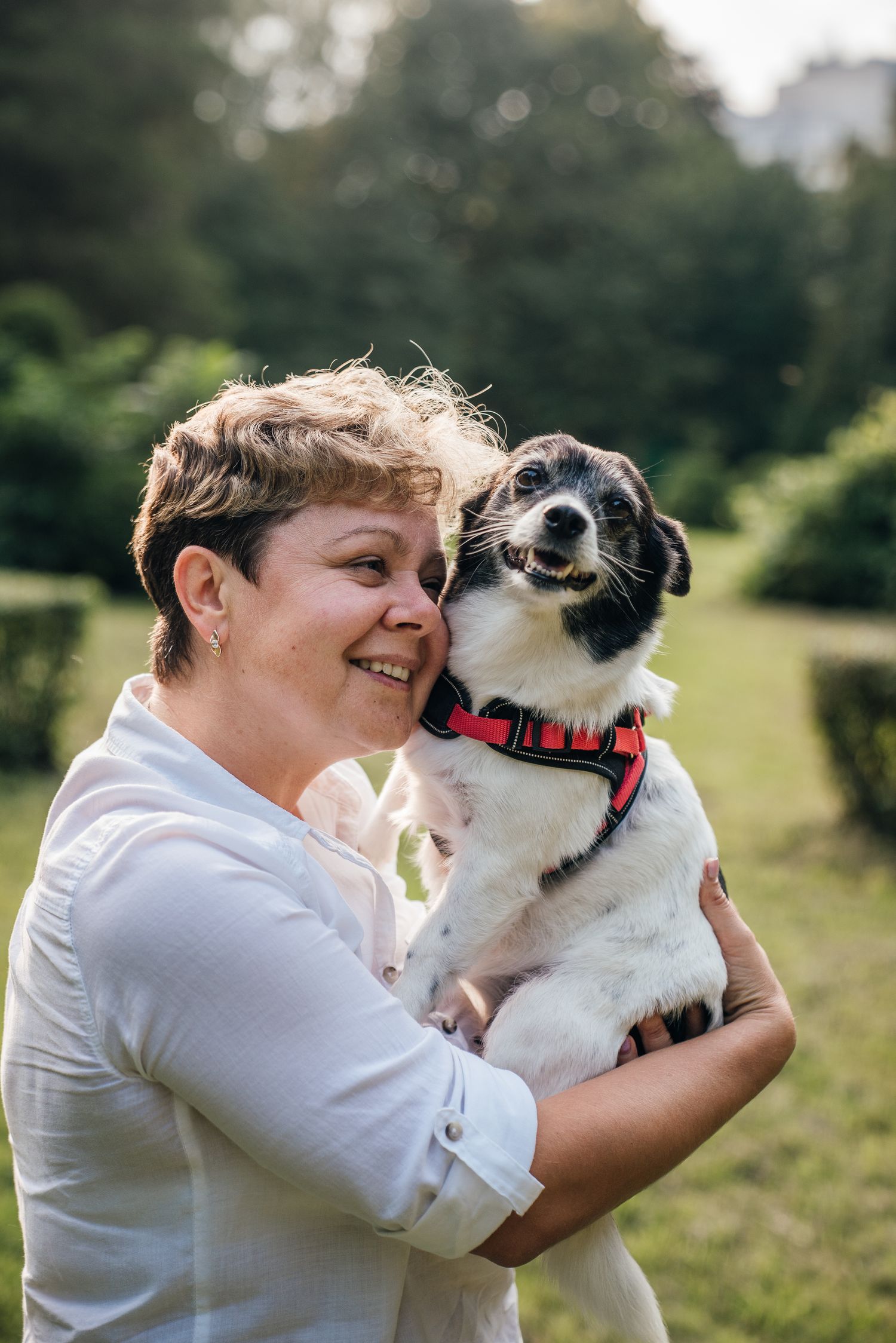 Smiling woman with short hair holding a small black and white dog in a red harness outdoors in a green park.