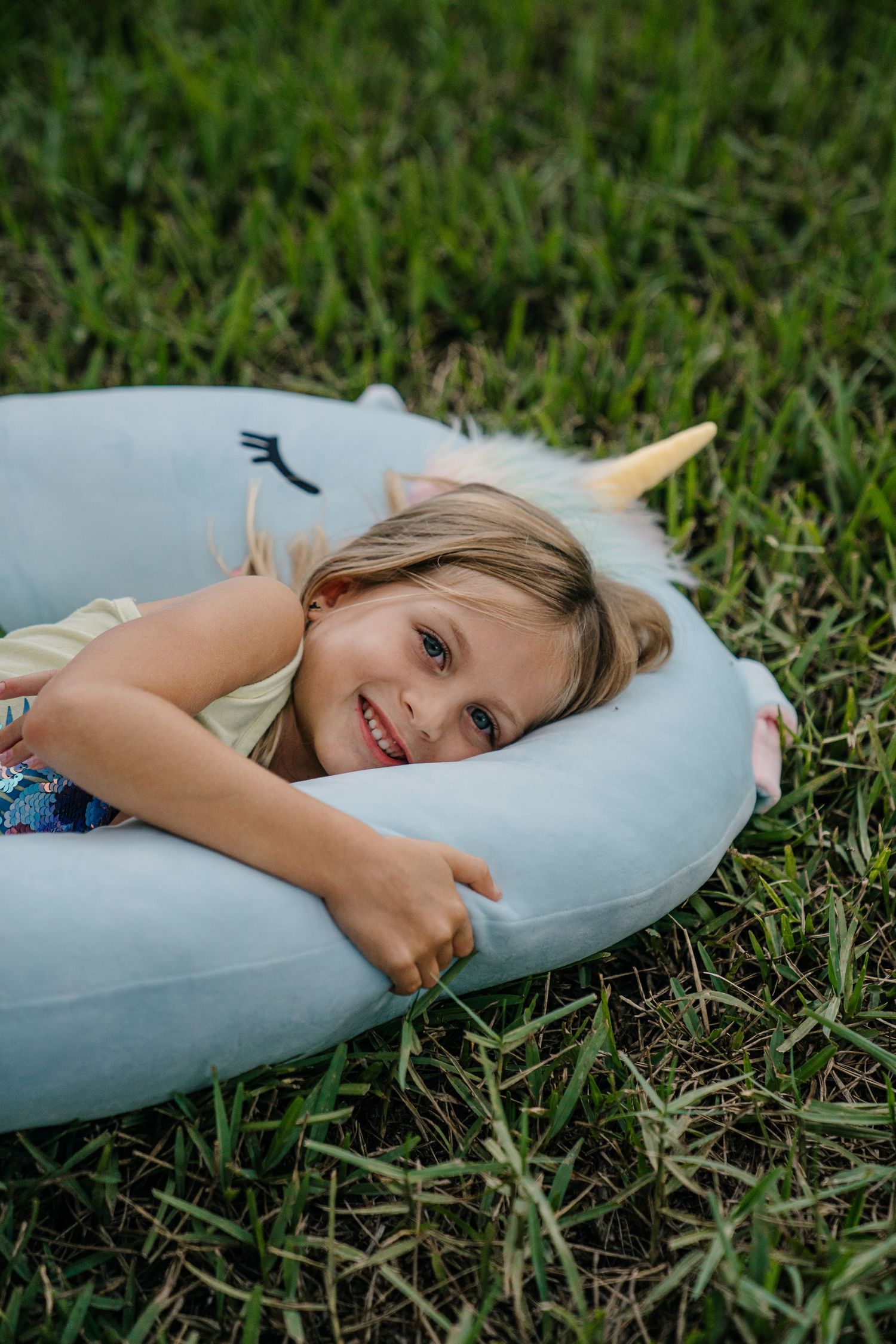 Young girl smiling while lying on an inflatable unicorn pool float on green grass outdoors.