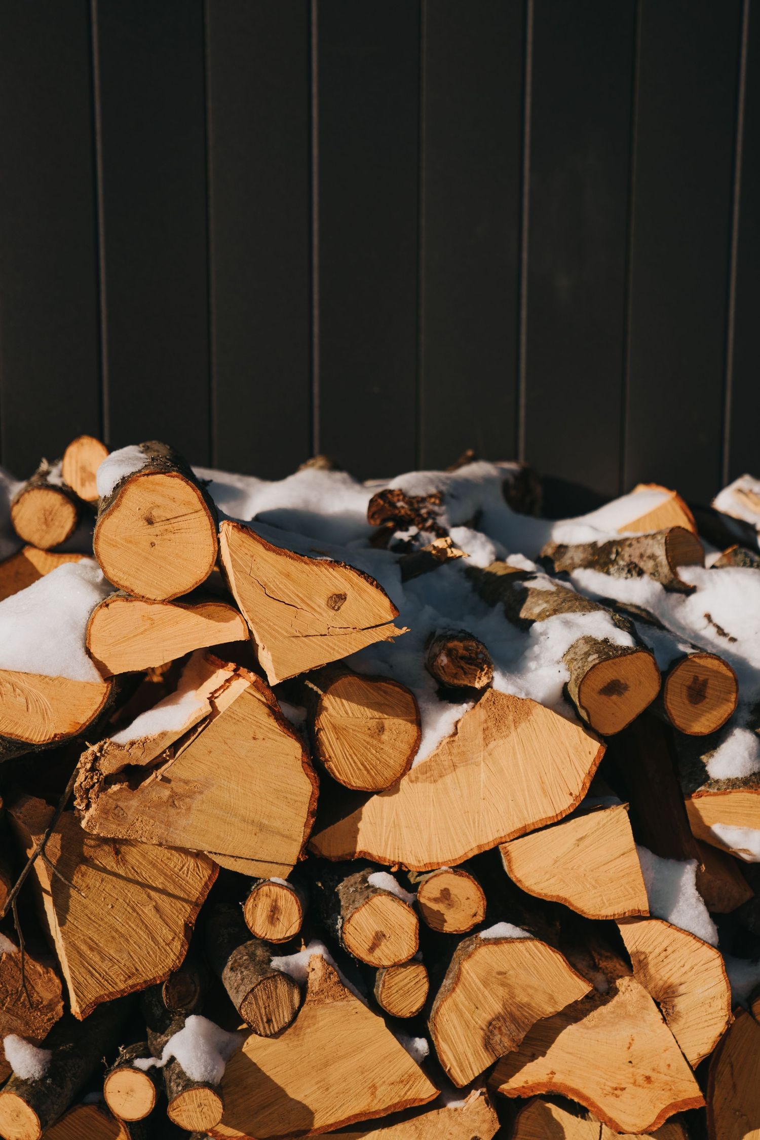 Stack of split firewood covered in snow against a dark wooden wall in winter sunlight.
