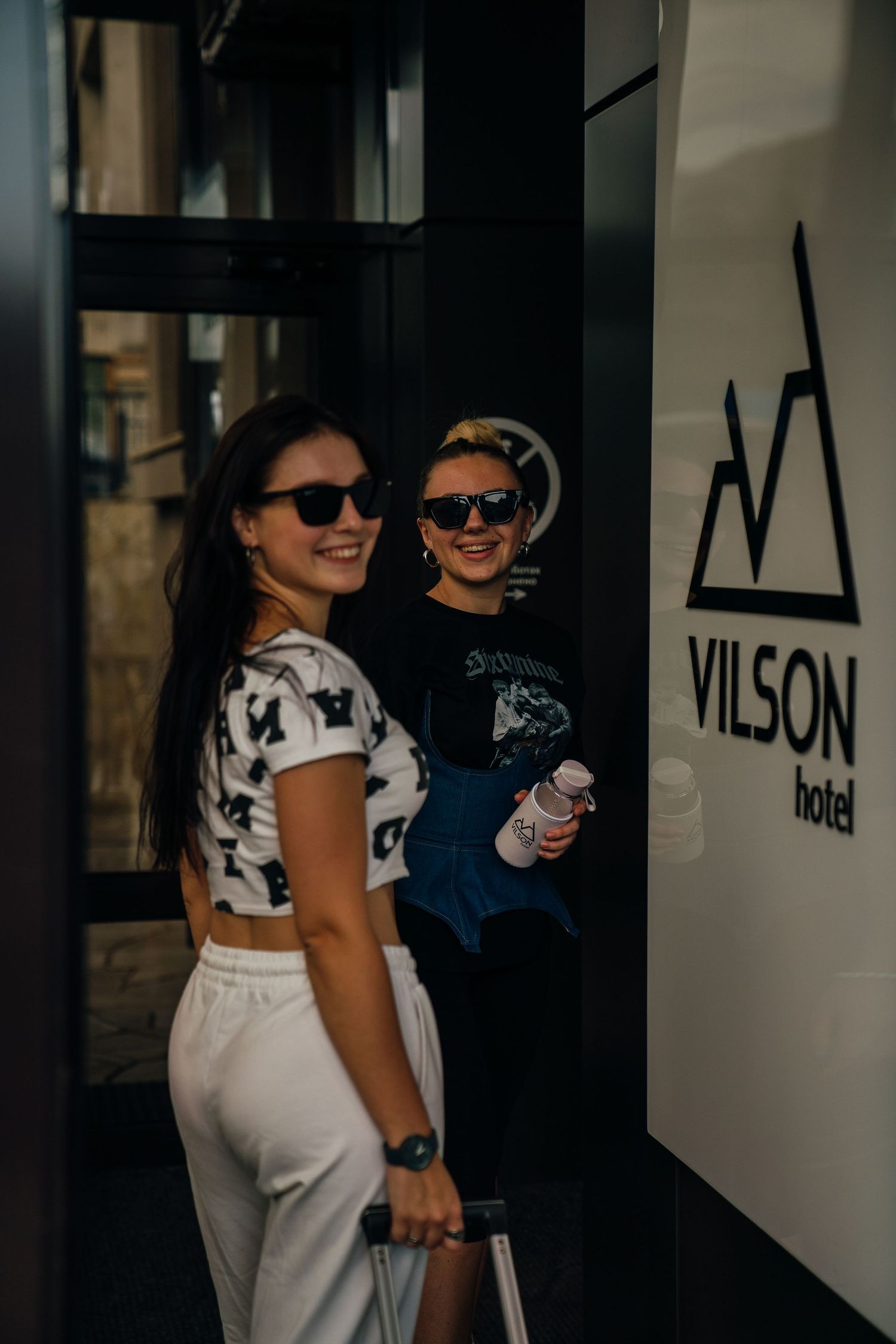Two smiling women wearing sunglasses arriving at Vilson Hotel entrance with luggage.
