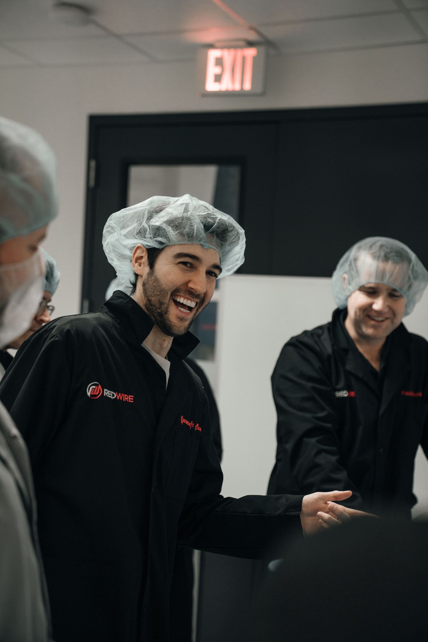 Two smiling workers in black uniforms and hairnets stand near an exit sign in a food production facility.