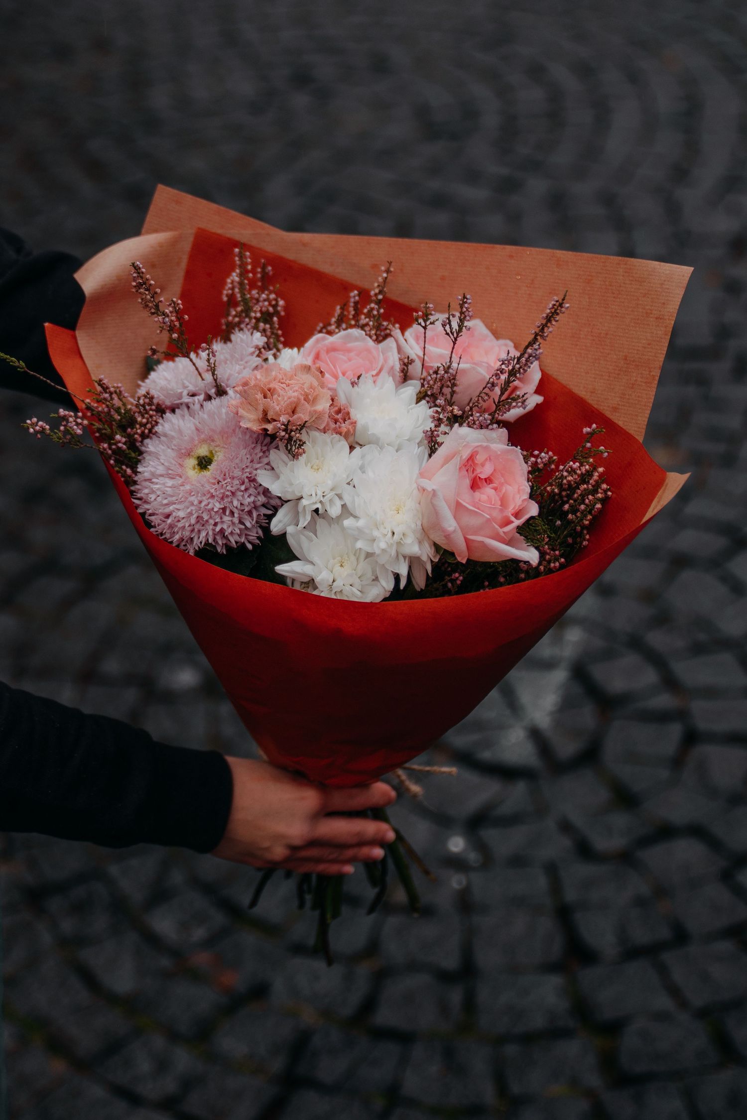 Hand holding a romantic bouquet of pink roses, white chrysanthemums, and dried flowers wrapped in red and kraft paper.