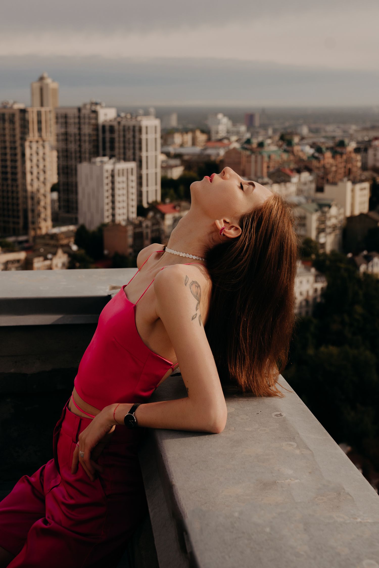 Young woman in pink outfit poses on rooftop balcony with city skyline backdrop at dusk.