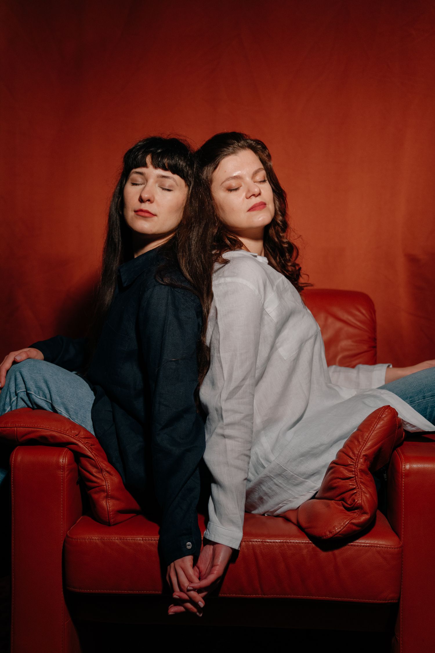 Two young women sitting back-to-back on a red leather sofa, eyes closed, holding hands against a red background.