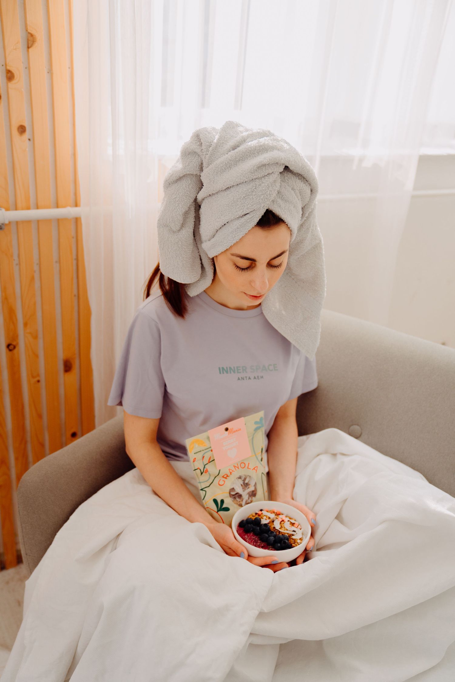 A woman with a towel on her head holding a bowl of granola with berries while relaxing on a sofa.