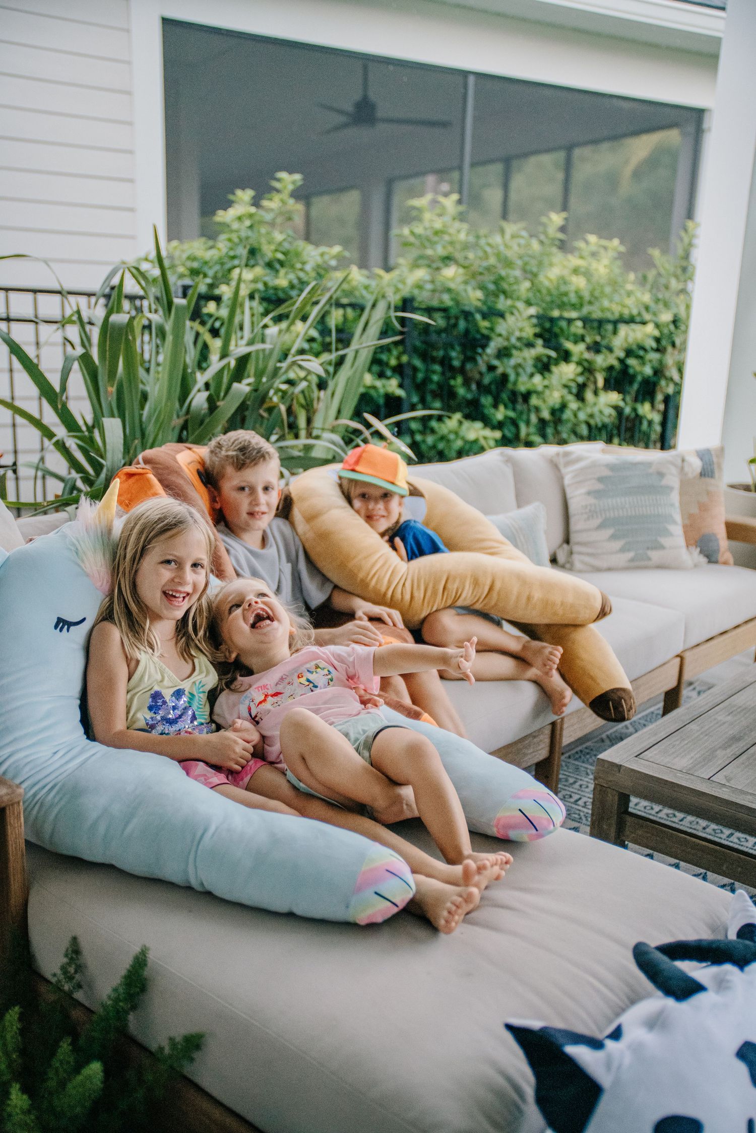 Four children laughing and playing on outdoor sofa with large unicorn stuffed animal pillow.