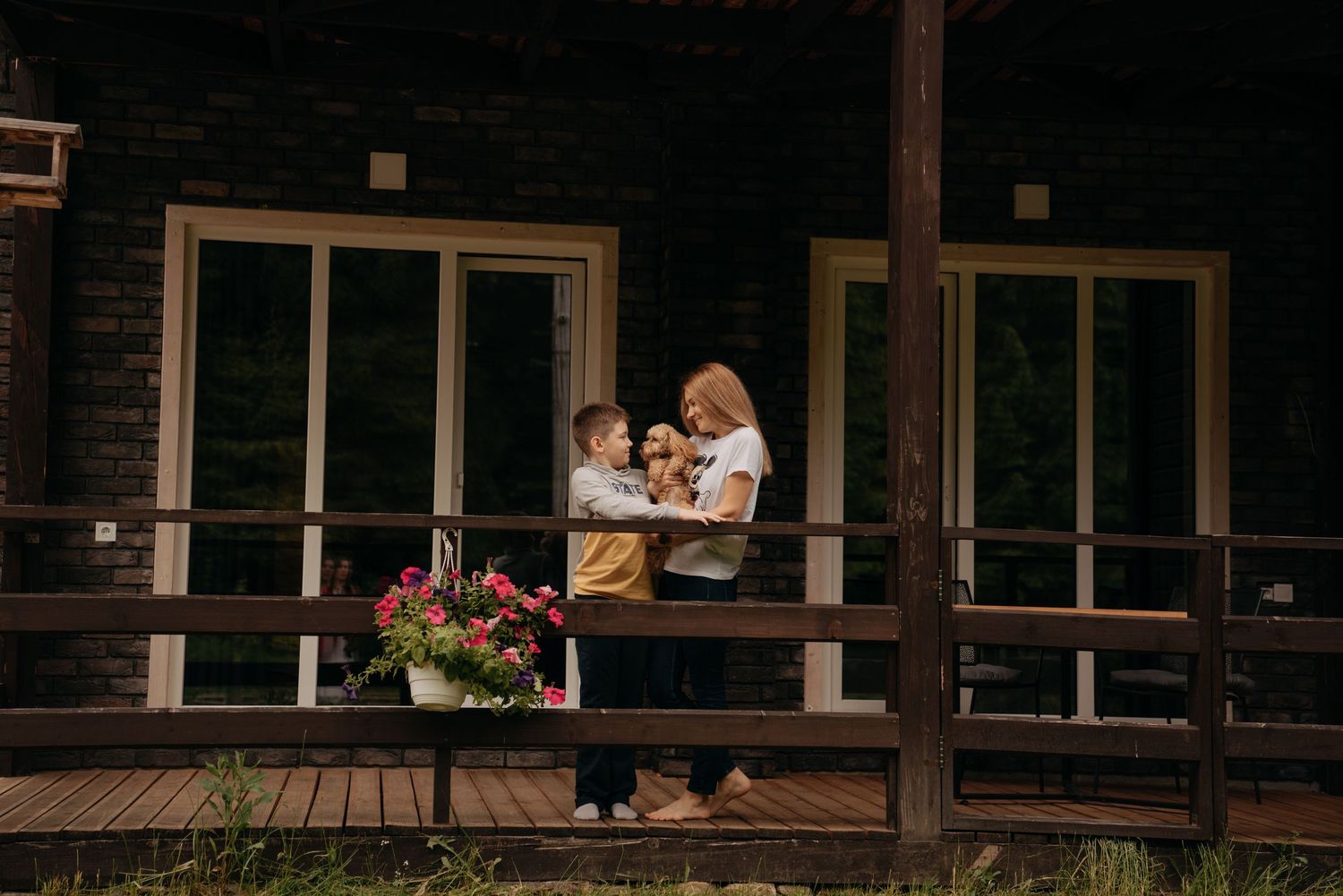 Family with child and dog enjoying time on wooden porch of dark log cabin home.