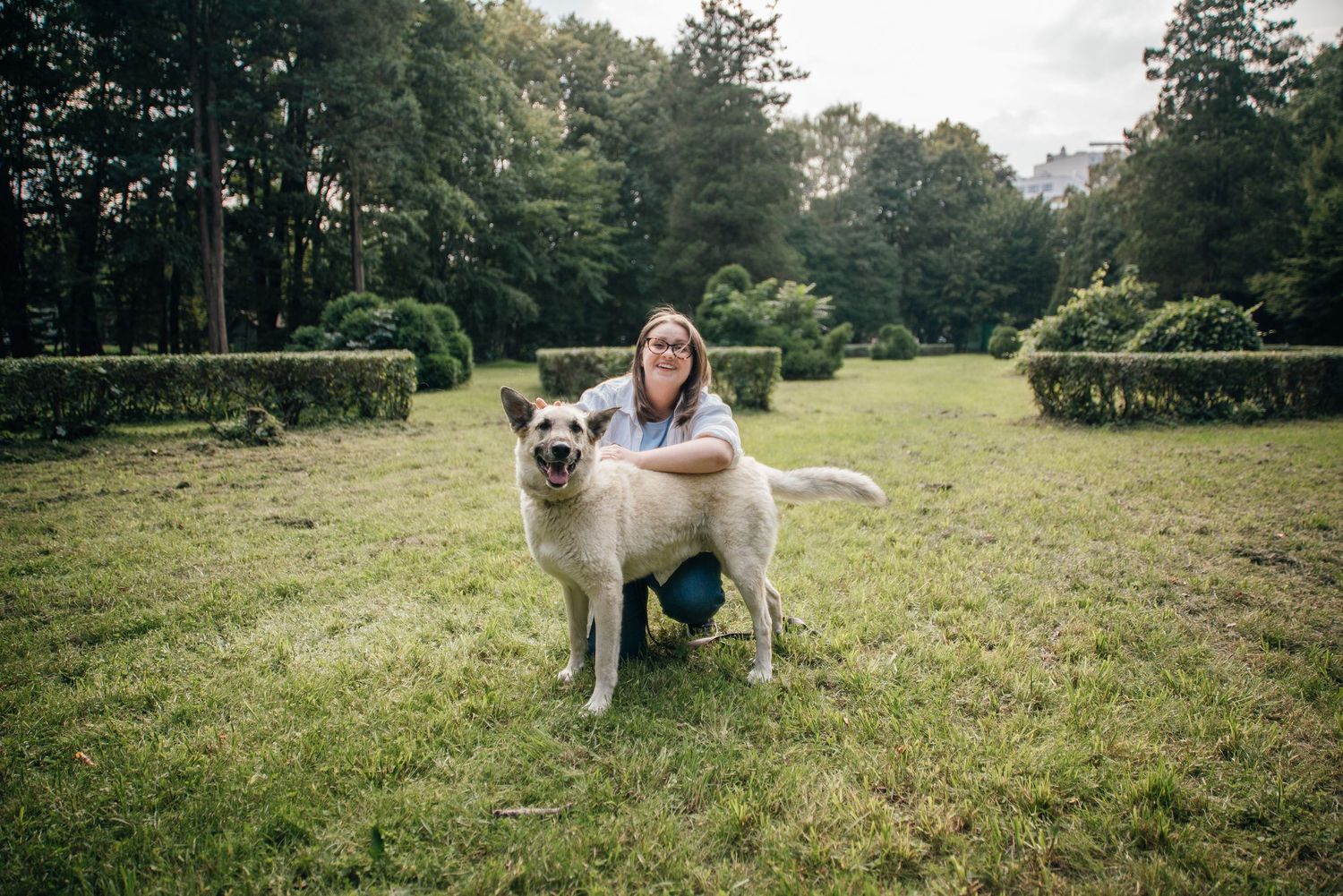 Woman kneeling beside a large white dog on a green lawn with manicured hedges and trees in the background.
