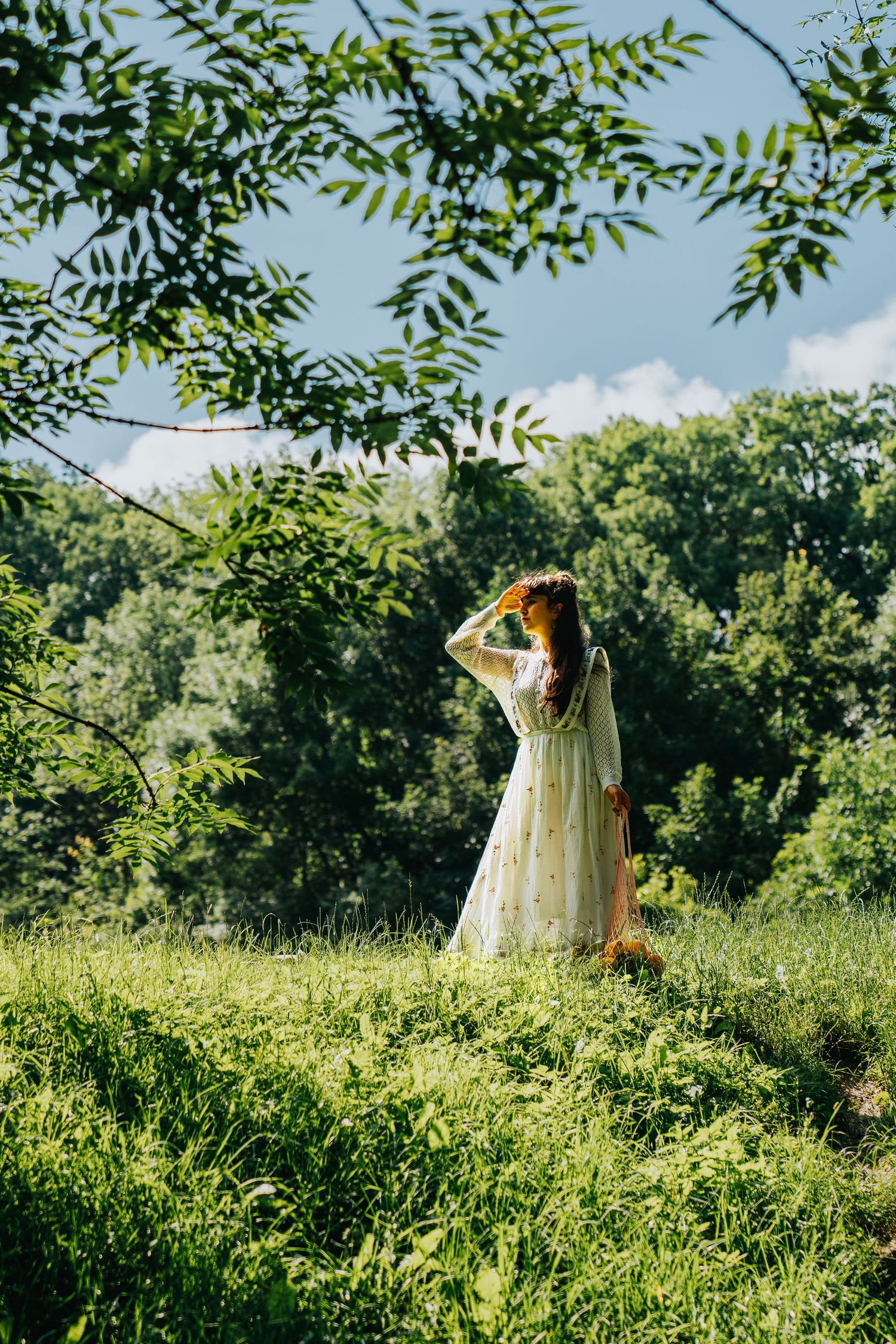 Woman in white floral dress standing in lush green meadow surrounded by trees on a sunny day.