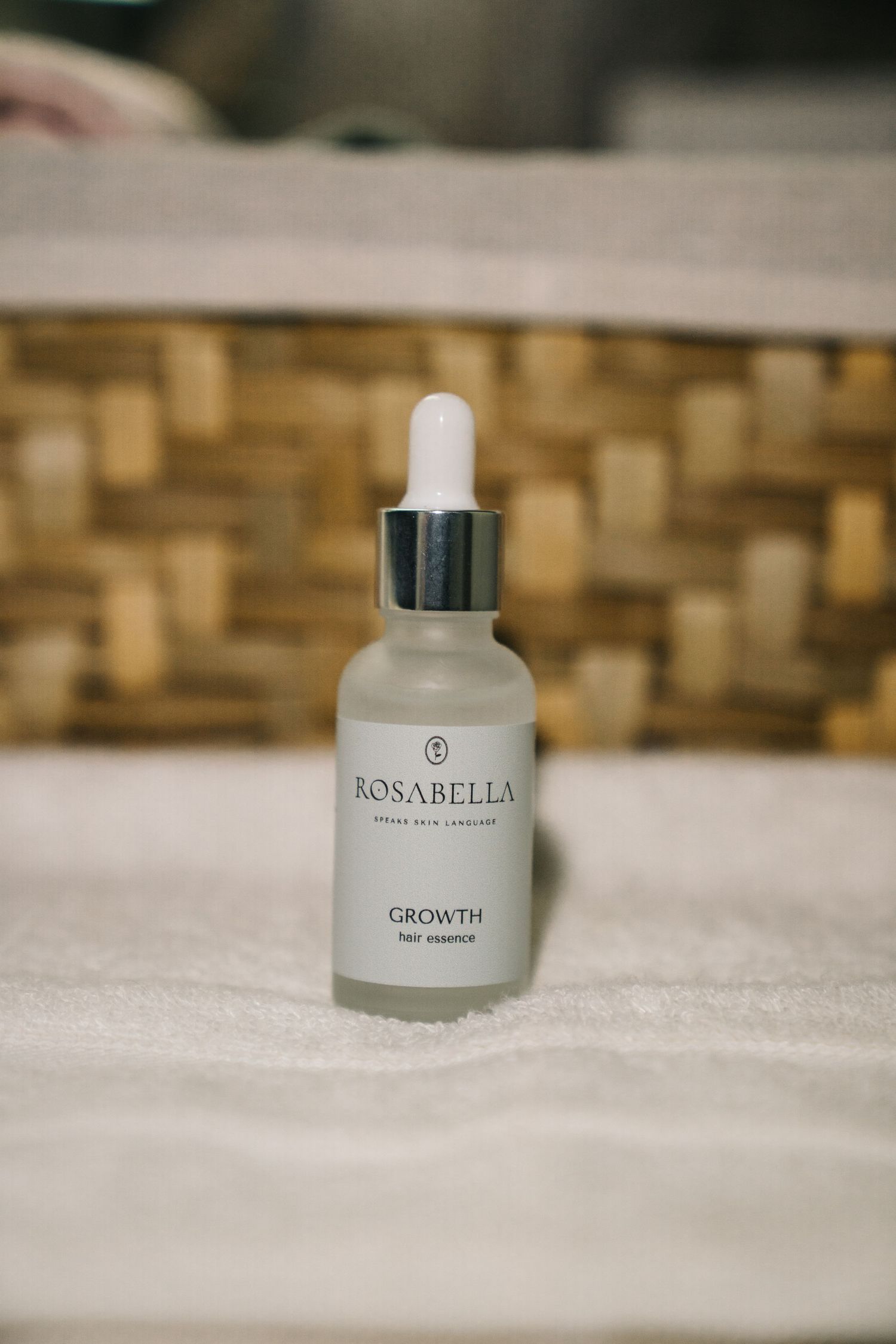 Rosabella Growth Hair Essence serum in frosted glass dropper bottle on white towel near wicker basket.