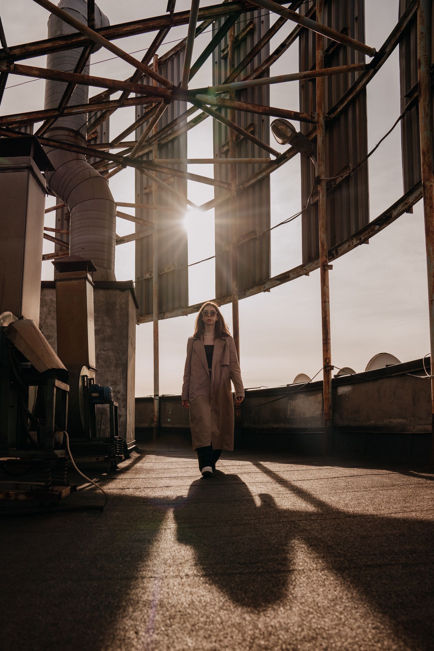 Woman in long coat walking on rooftop with sunlight streaming through industrial glass windows casting dramatic shadows.