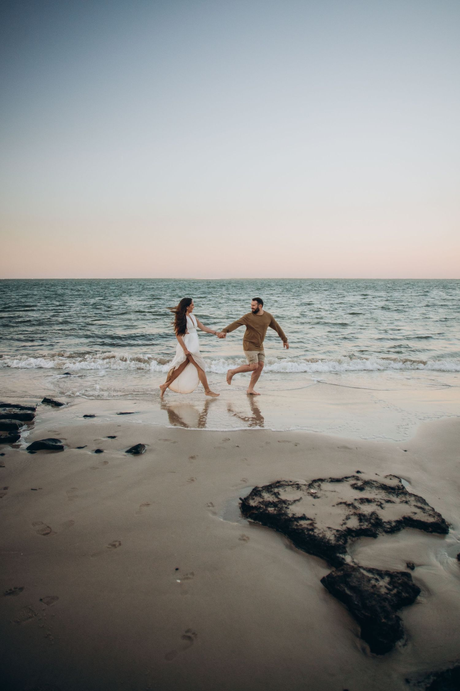 Couple running playfully along the beach at sunset with dark rocks in foreground.