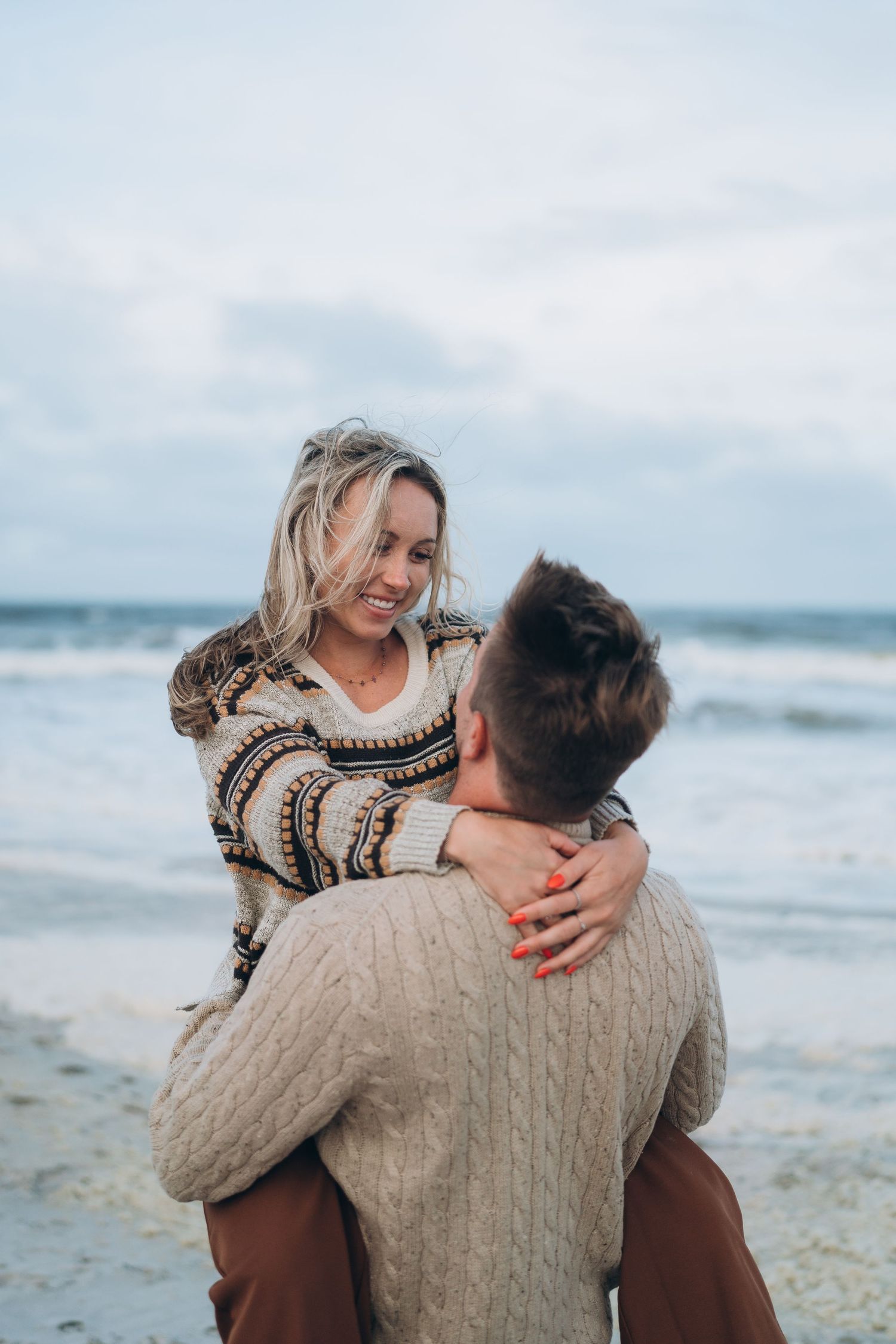 A couple embraces while wearing cozy sweaters on a windswept beach.