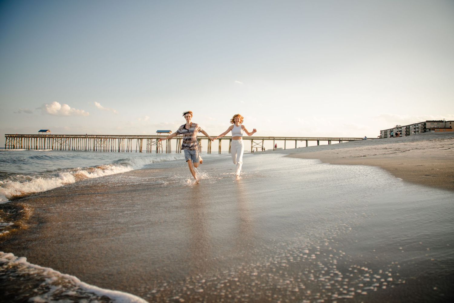 Wide angle beach scene with pier and figures silhouetted against setting sun.