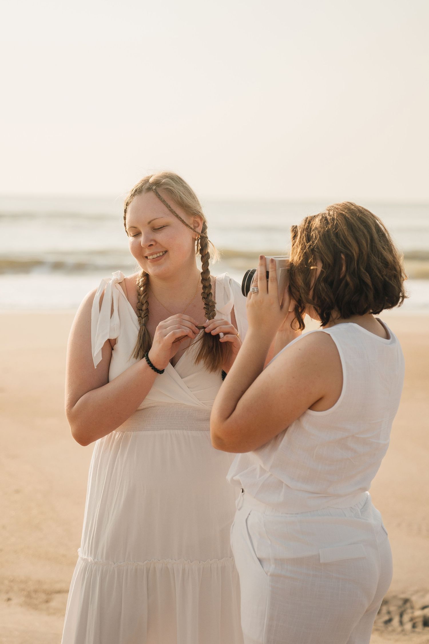 Two people in white dresses share a playful moment on a beach during sunset.