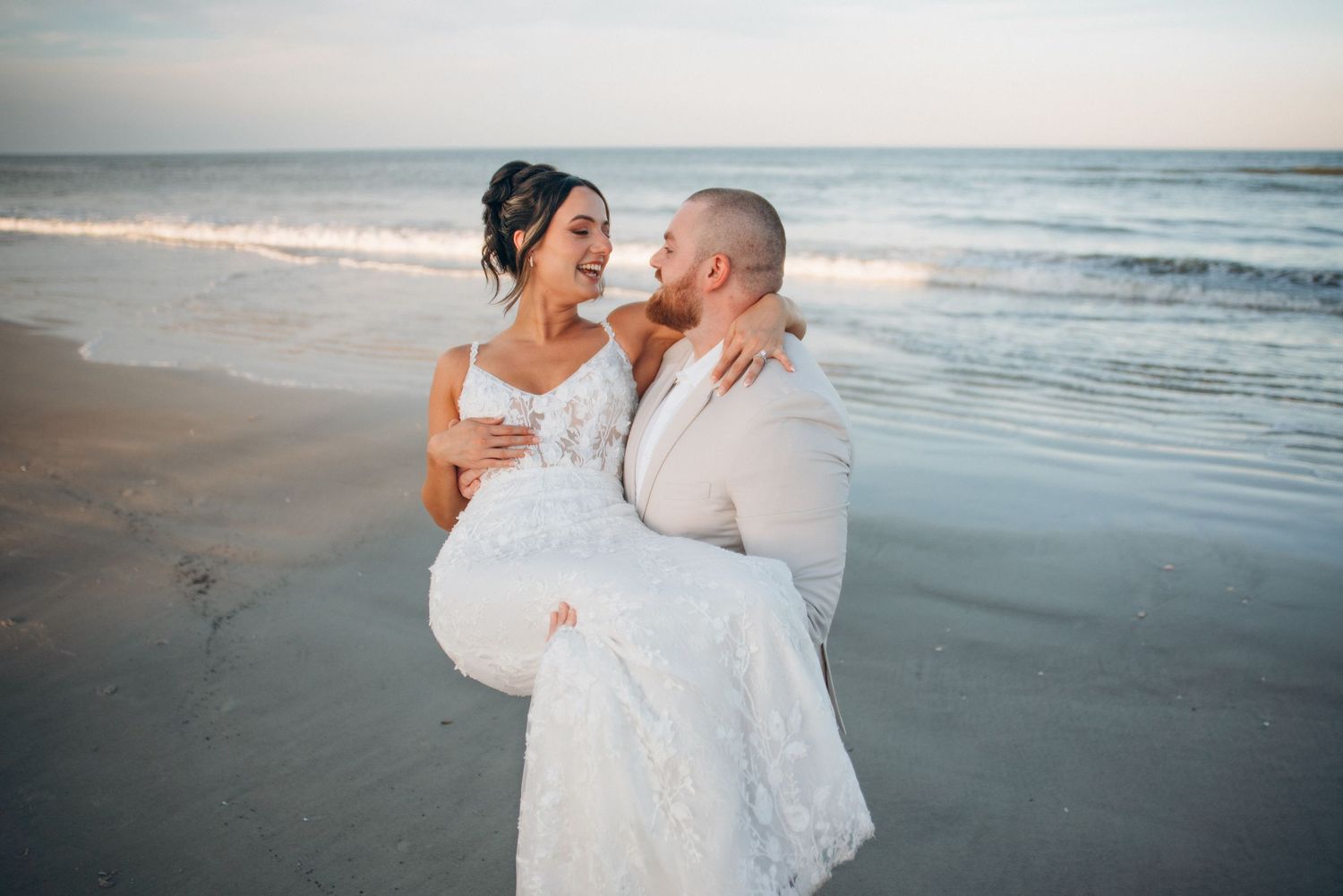 Groom carries bride in white lace gown on sandy beach at sunset with ocean waves in background.