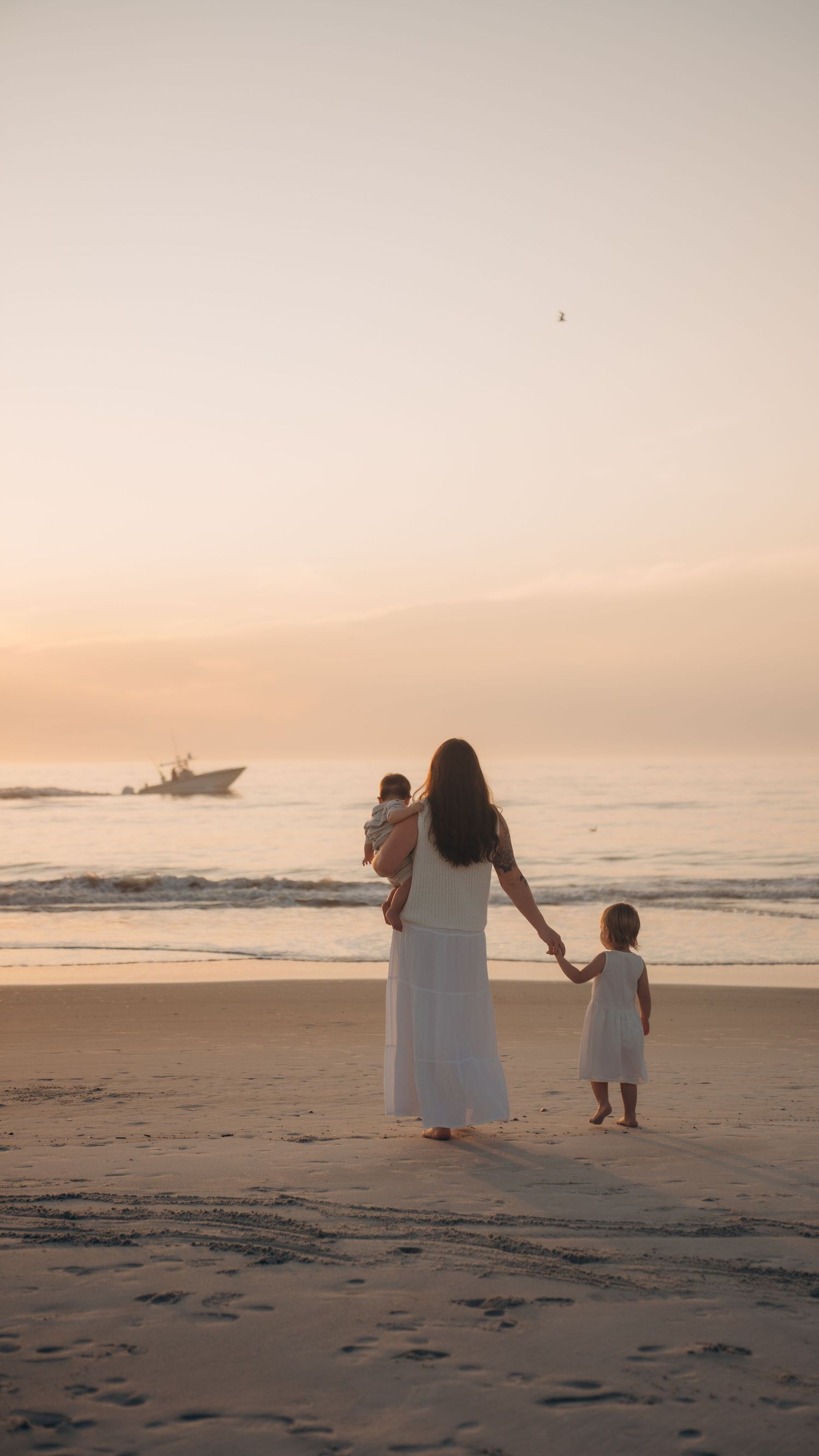 A mother in a white dress holds her baby and toddler's hand while watching a serene ocean sunset on a sandy beach.
