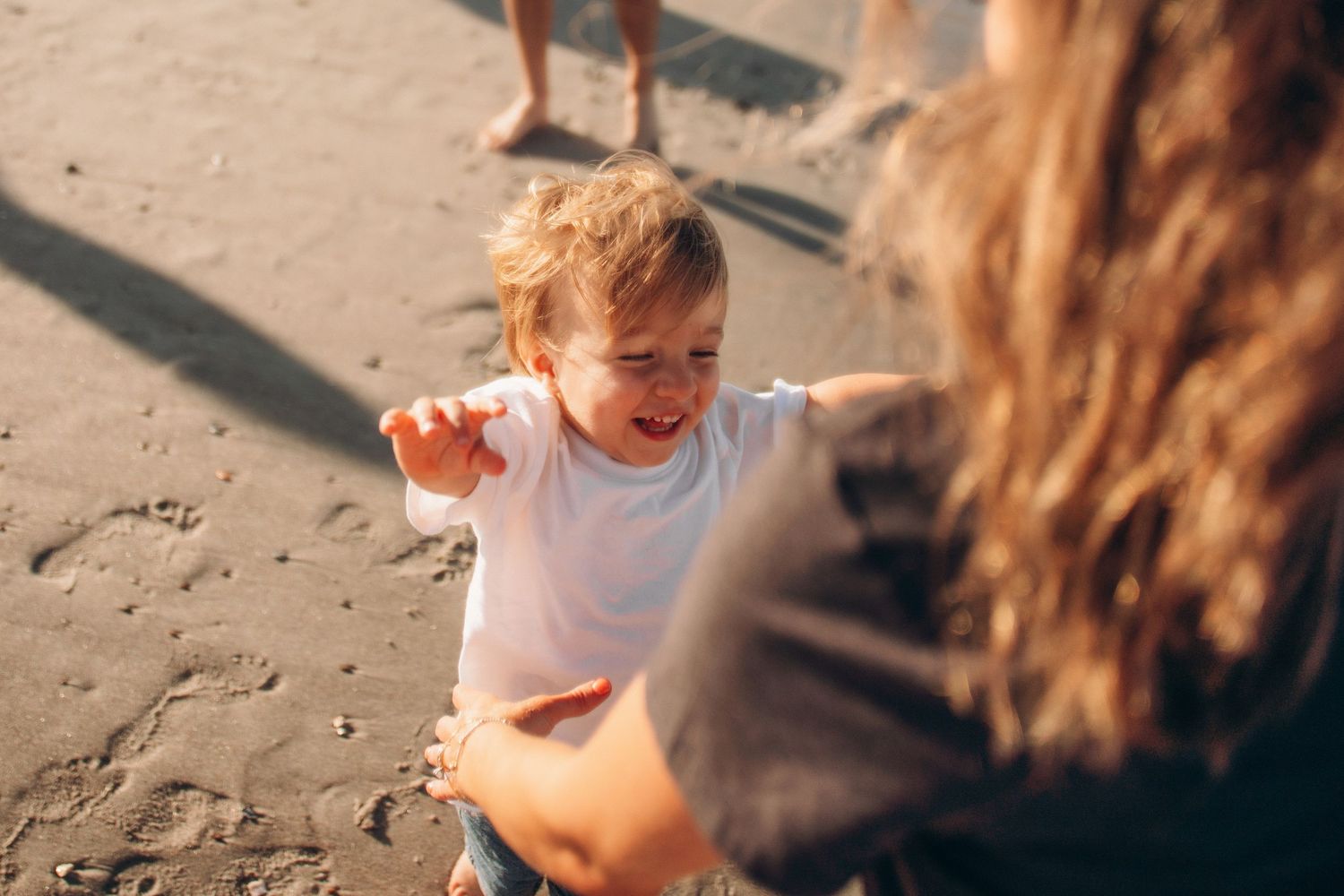 Toddler laughing and reaching out on a sandy beach while being held by a woman in warm sunlight.