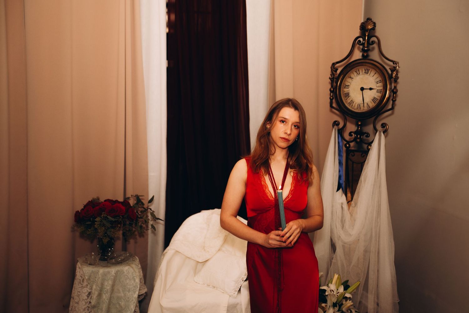 Woman in red dress holding fabric near ornate clock and curtains in elegant room.