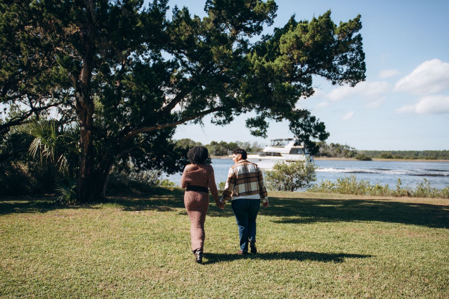 A couple walks together on a grassy waterfront path beneath towering trees on a sunny afternoon.