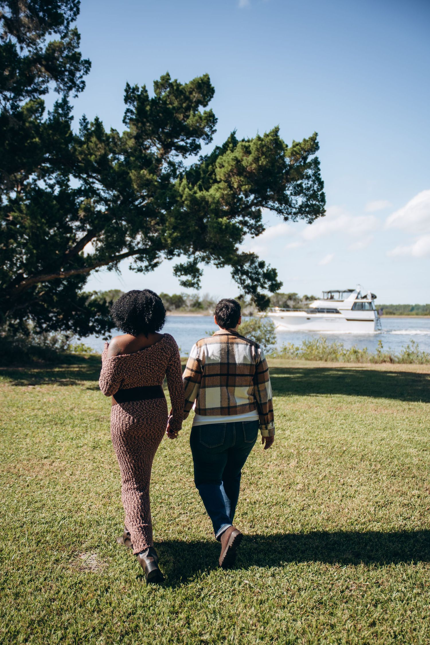 Two people walk together across a grassy hill overlooking a waterfront on a sunny day.