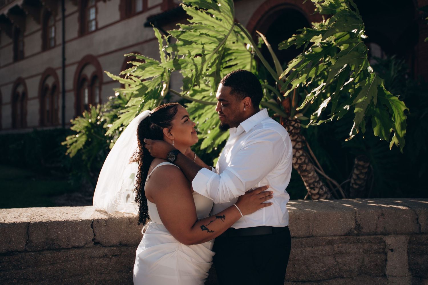 A bride and groom share a romantic embrace outdoors near a stone wall with lush greenery on their wedding day.