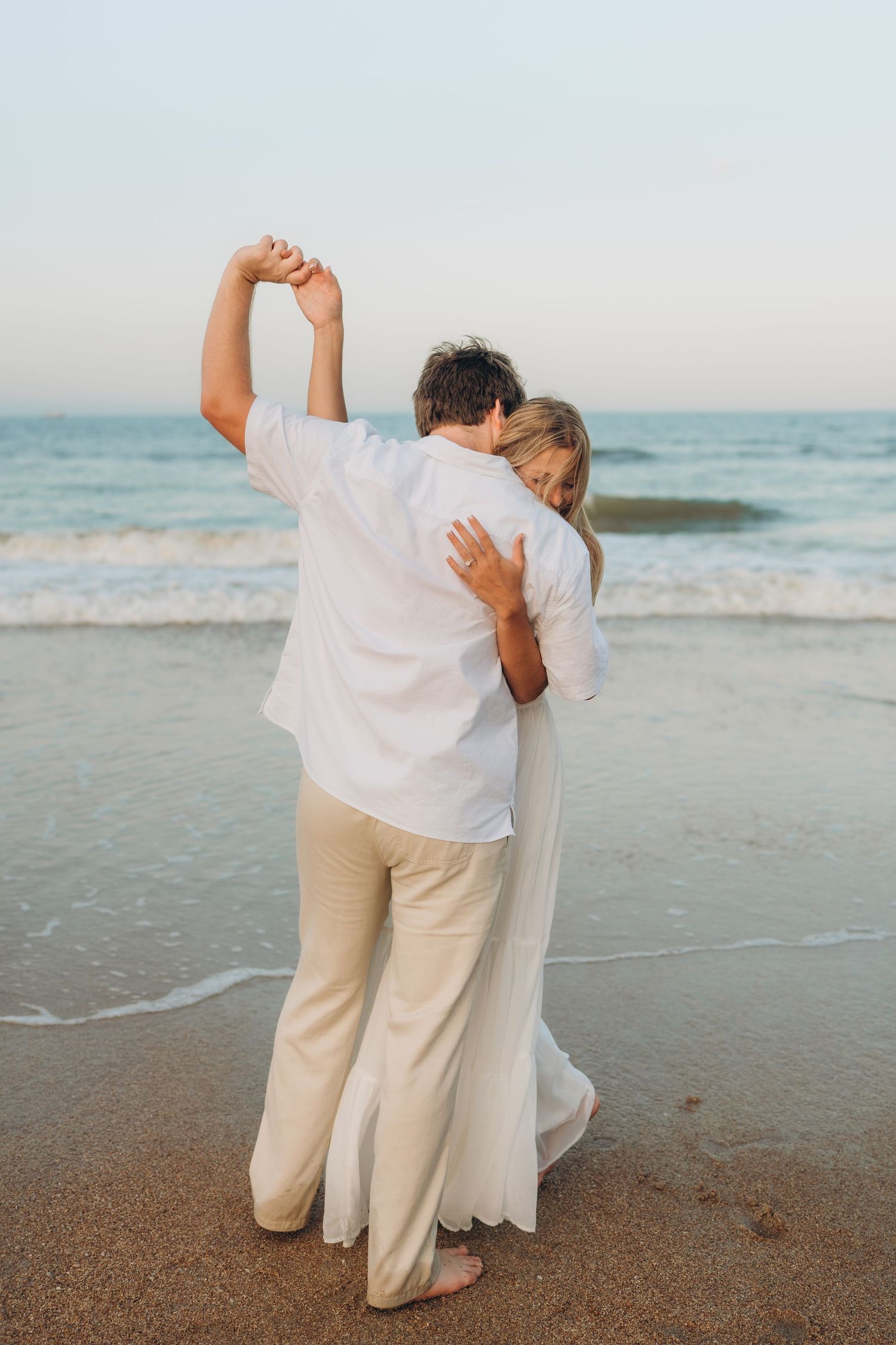 A couple dressed in white embraces while standing by the ocean waves at sunset.