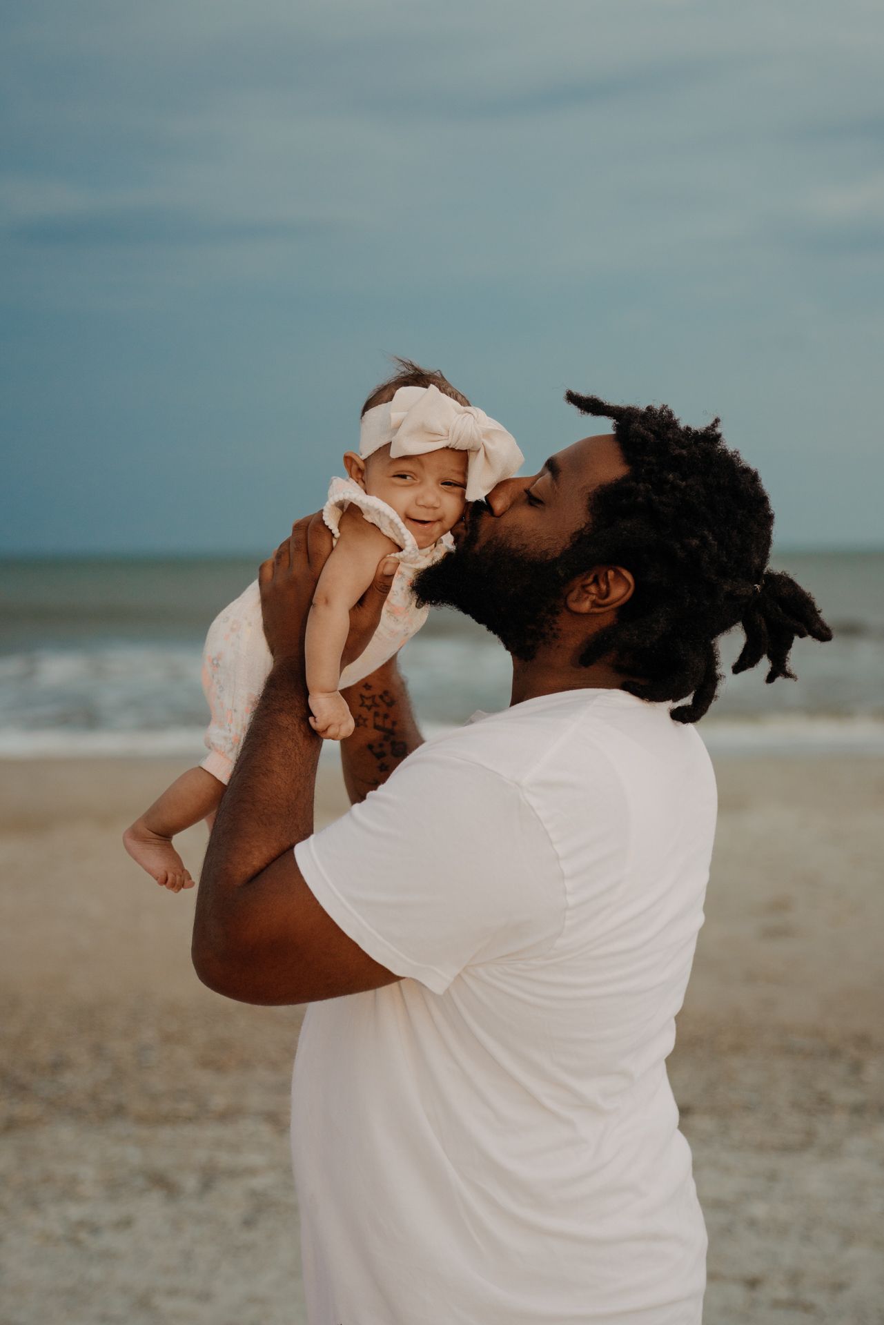 Parent lifting and kissing baby on a peaceful beach at dusk.