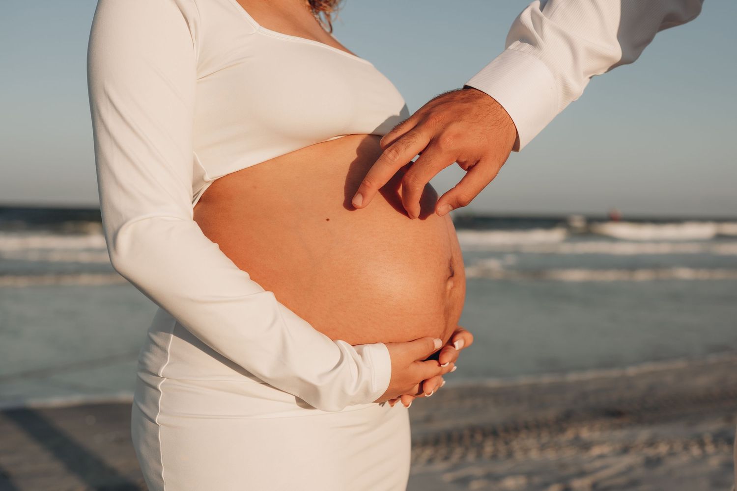 Intimate beach maternity photos showing tender moments between a couple in white attire.