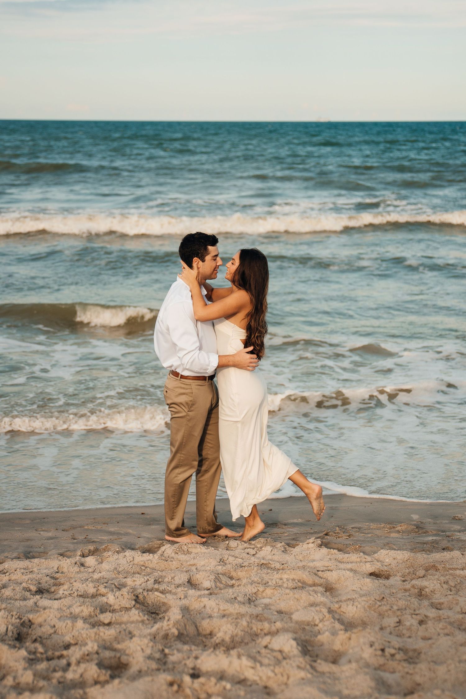 A joyful couple embraces while standing on the beach with ocean waves behind them.