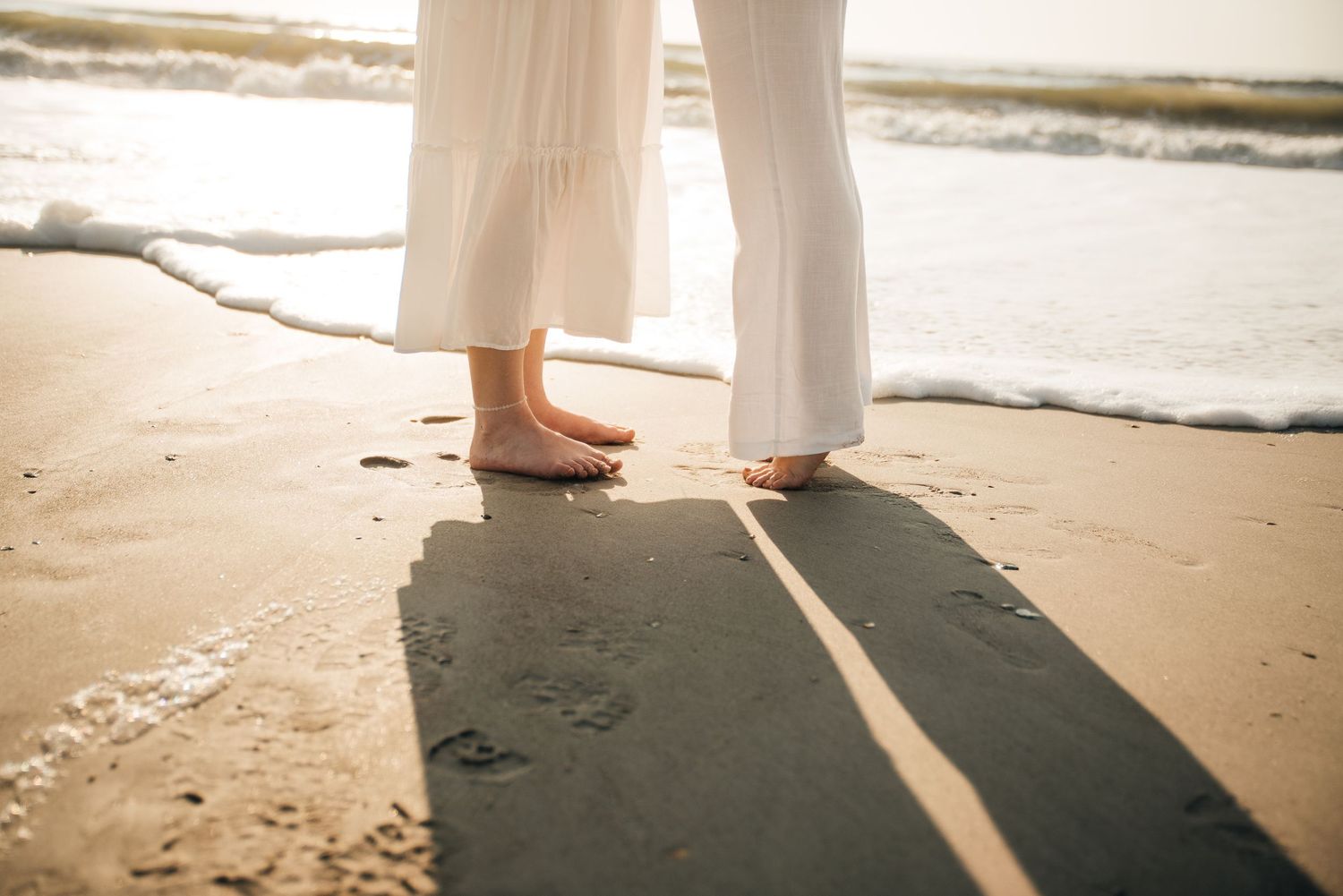 Footprints in the sand as a couple walks along the beach in white wedding attire.