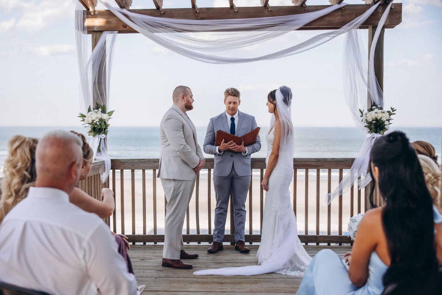 Outdoor beach wedding ceremony on a wooden deck with bride, groom, and officiant under a white draped arbor.