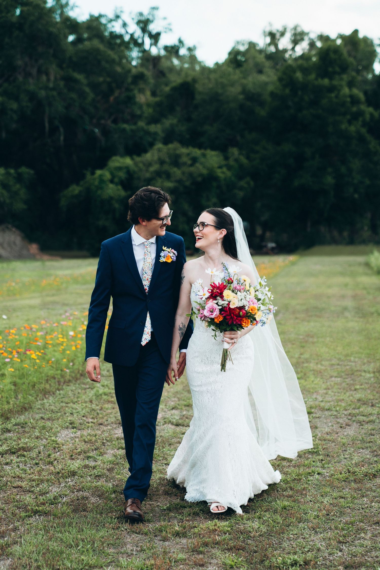 A bride and groom walk hand-in-hand through a green meadow, she in lace gown with veil, he in navy suit with floral boutonniere.