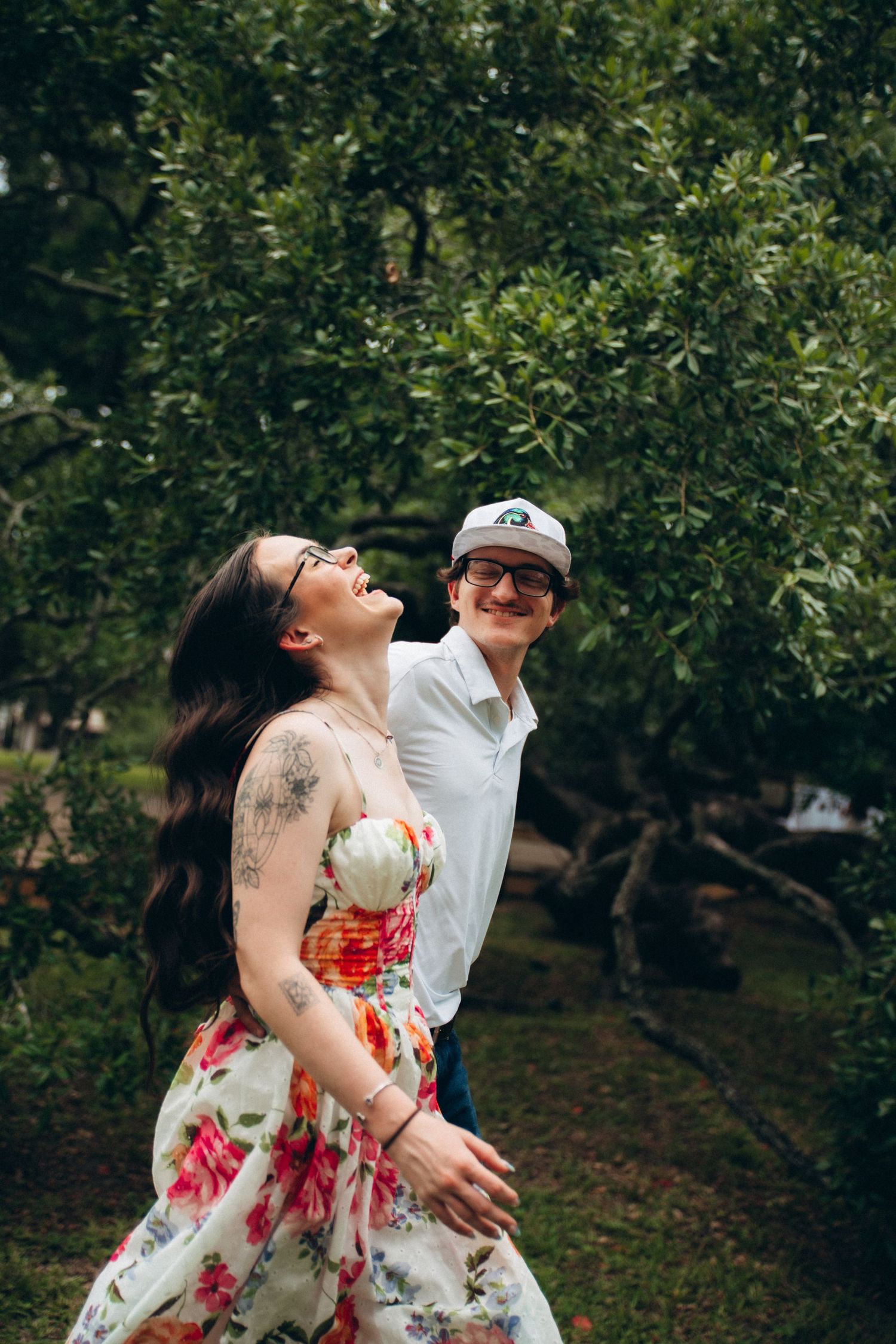 Laughing couple embracing near lush green trees, woman in floral dress with visible arm tattoo.
