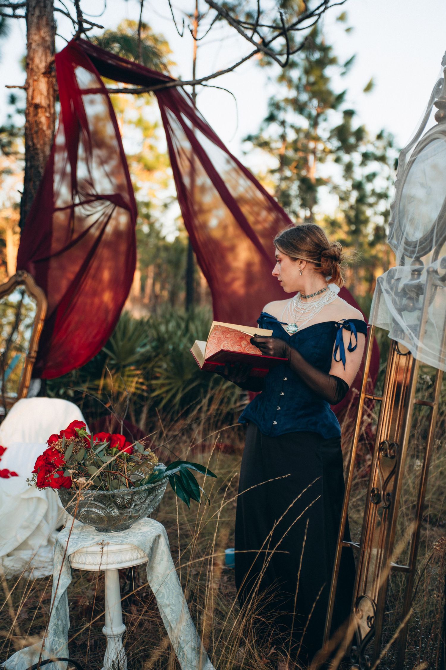 A woman in a Victorian navy dress reads a book outdoors among red roses and flowing crimson fabric in a forest setting.